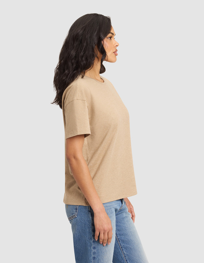 A woman with wavy dark hair stands in profile, wearing Cozy Earth's Women's All Day Tee in beige and blue jeans, set against a plain light gray background.