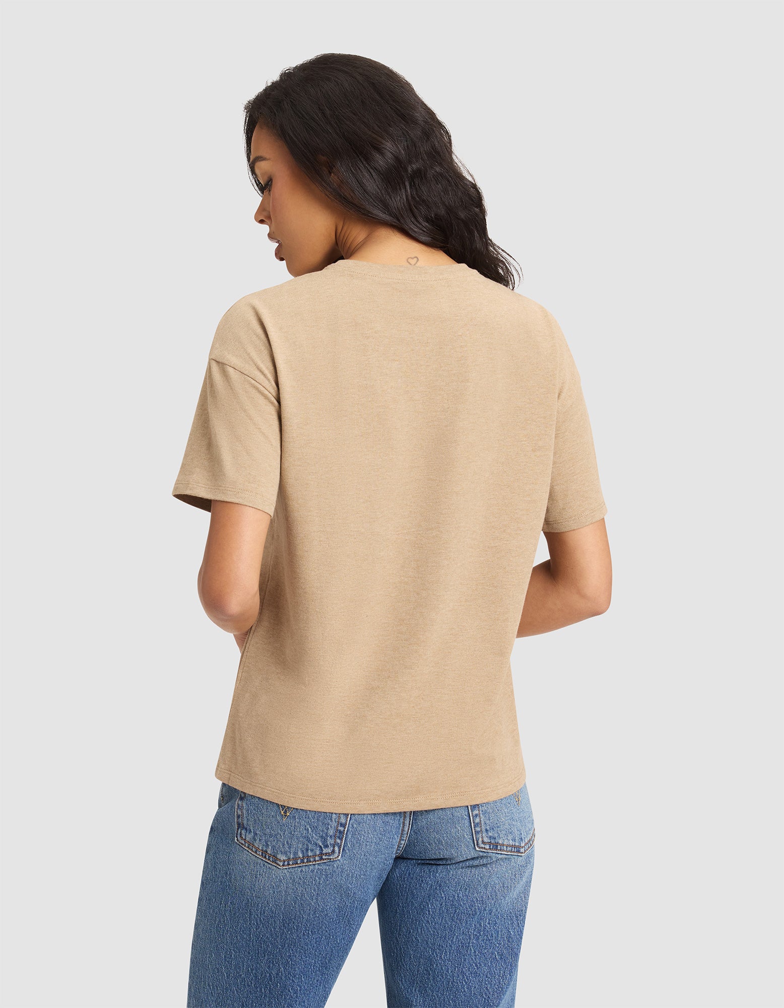 A woman with long dark hair is shown from the back against a plain light background, wearing the Cozy Earth Women's All Day Tee in beige and blue jeans. 