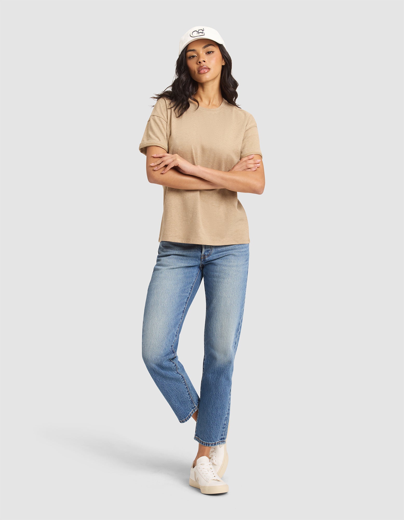 A woman stands against a plain background, arms crossed and one leg slightly bent, wearing Cozy Earth’s Women's All Day Tee in beige, paired with blue jeans, white sneakers, and a white baseball cap. 