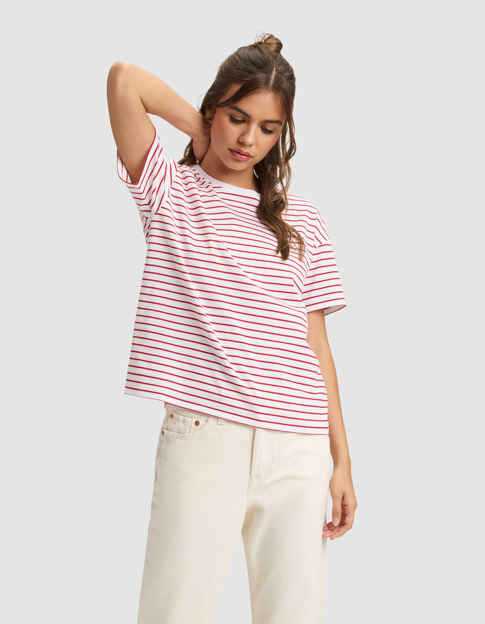 A woman models the Cozy Earth Women's All Day Tee, a white t-shirt with red stripes, paired with off-white pants. She stands with one hand behind her head, looking down against a plain light gray background.