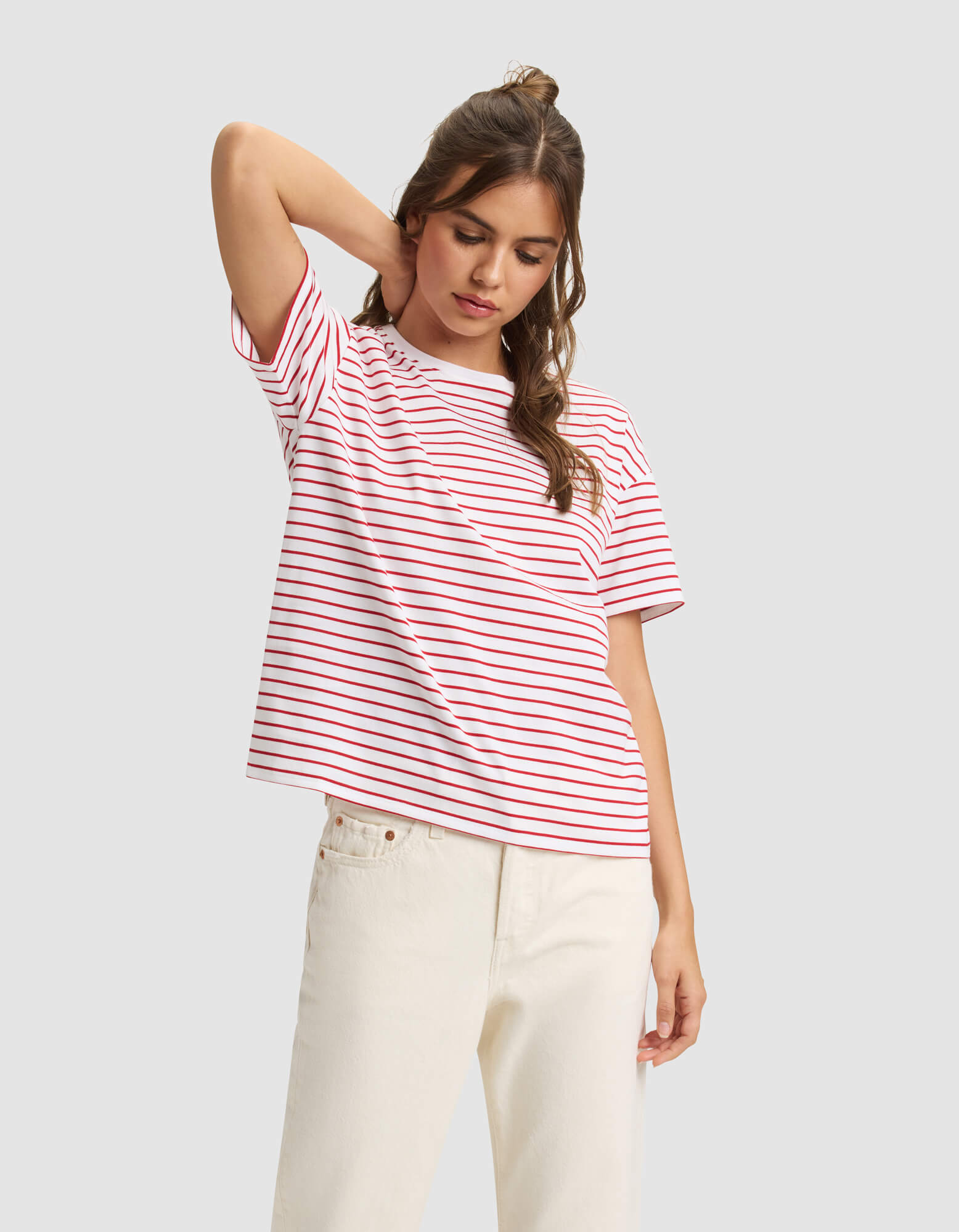 A woman models the Cozy Earth Women's All Day Tee, a white t-shirt with red stripes, paired with off-white pants. She stands with one hand behind her head, looking down against a plain light gray background. 