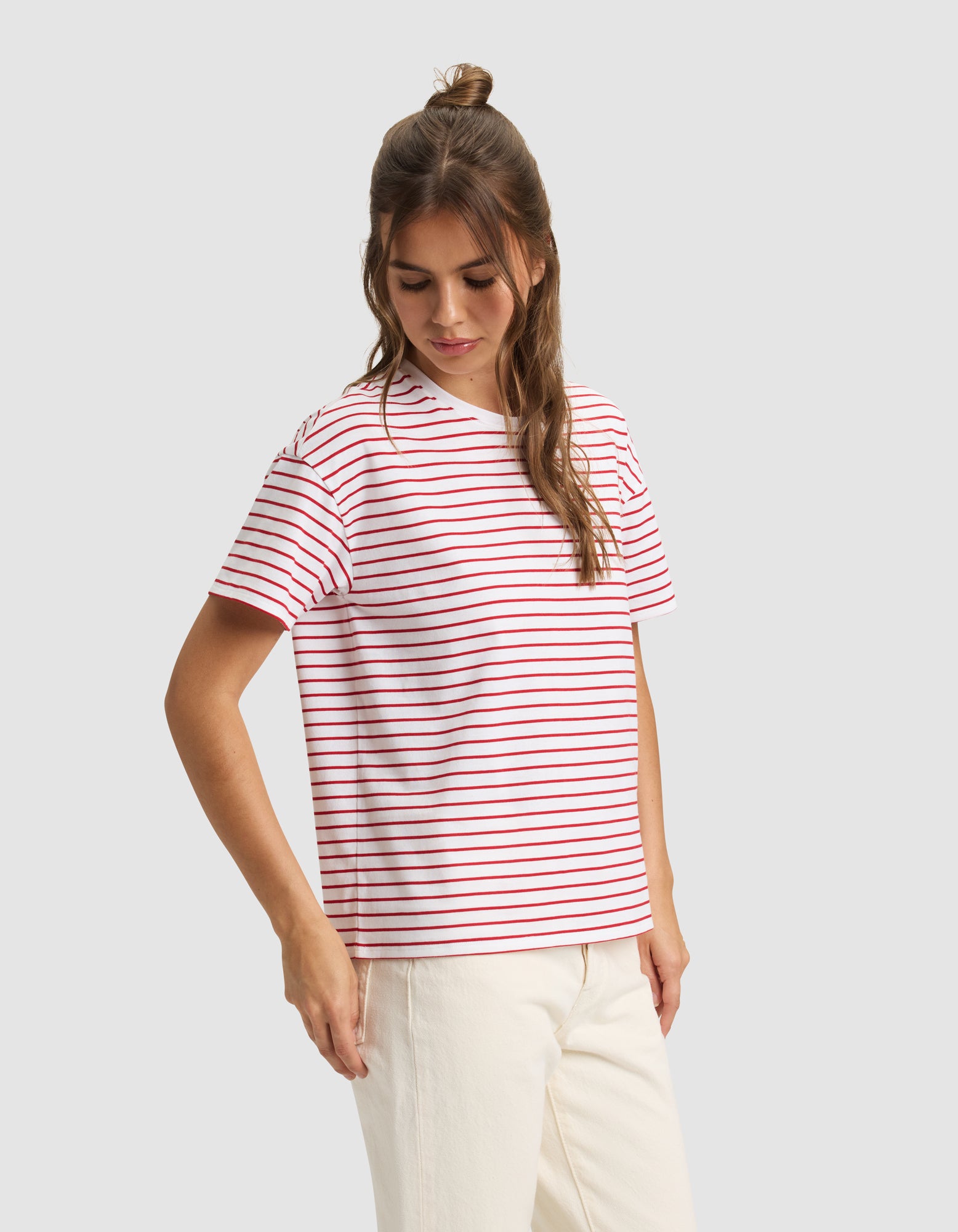 A woman models the Cozy Earth Women's All Day Tee—a white T-shirt with red and blue stripes—paired with off-white pants, standing against a light gray background.