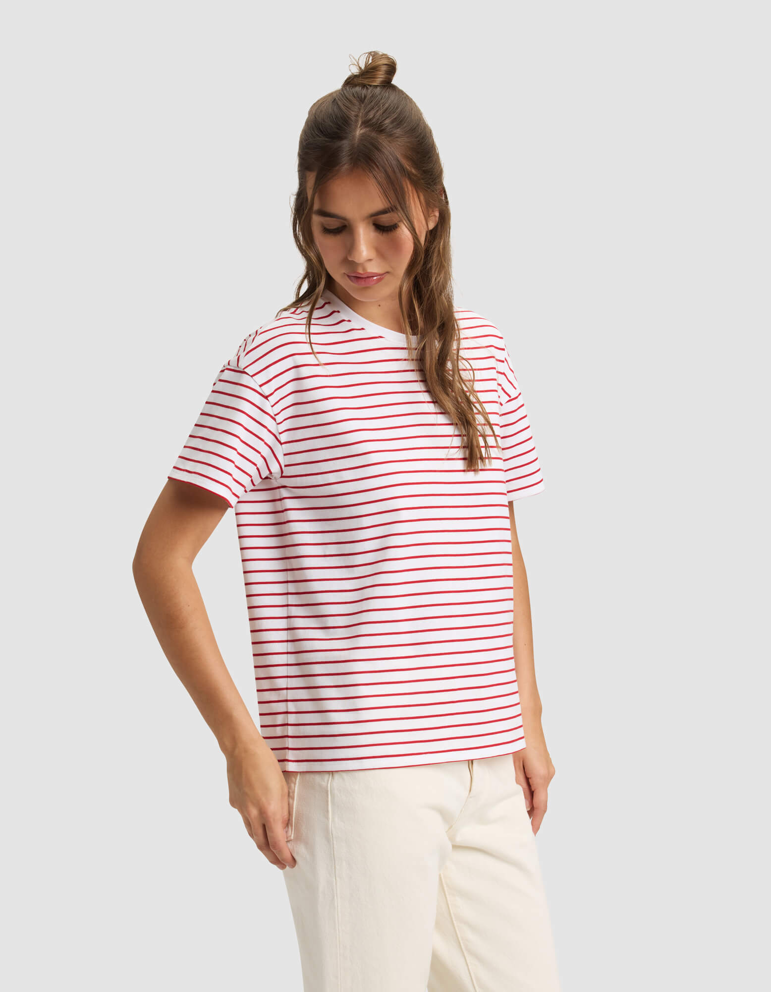 A woman models the Cozy Earth Women's All Day Tee—a white T-shirt with red and blue stripes—paired with off-white pants, standing against a light gray background. 