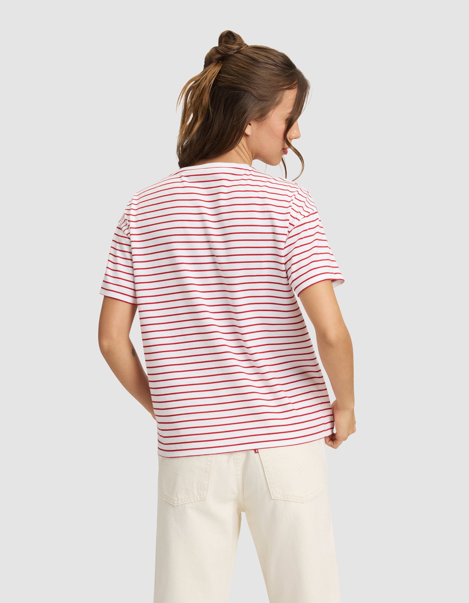 A woman with light brown hair in a half-up style is shown from the back wearing Cozy Earth’s Women’s All Day Tee—white with red and blue stripes—and off-white pants, standing against a plain light gray background.