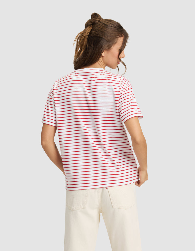 A woman with light brown hair in a half-up style is shown from the back wearing Cozy Earth’s Women’s All Day Tee—white with red and blue stripes—and off-white pants, standing against a plain light gray background.
