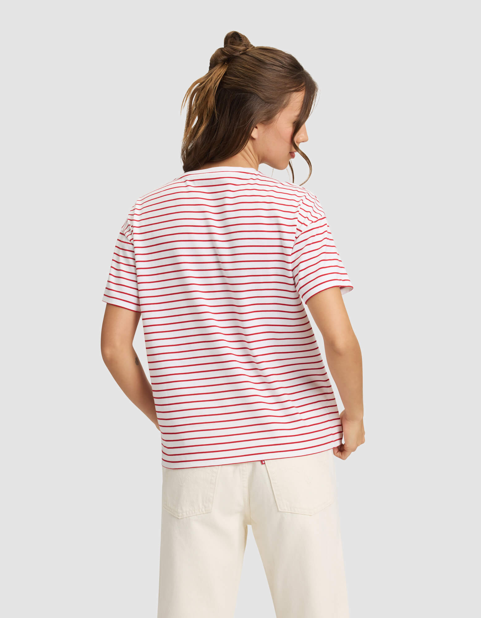 A woman with light brown hair in a half-up style is shown from the back wearing Cozy Earth’s Women’s All Day Tee—white with red and blue stripes—and off-white pants, standing against a plain light gray background. 