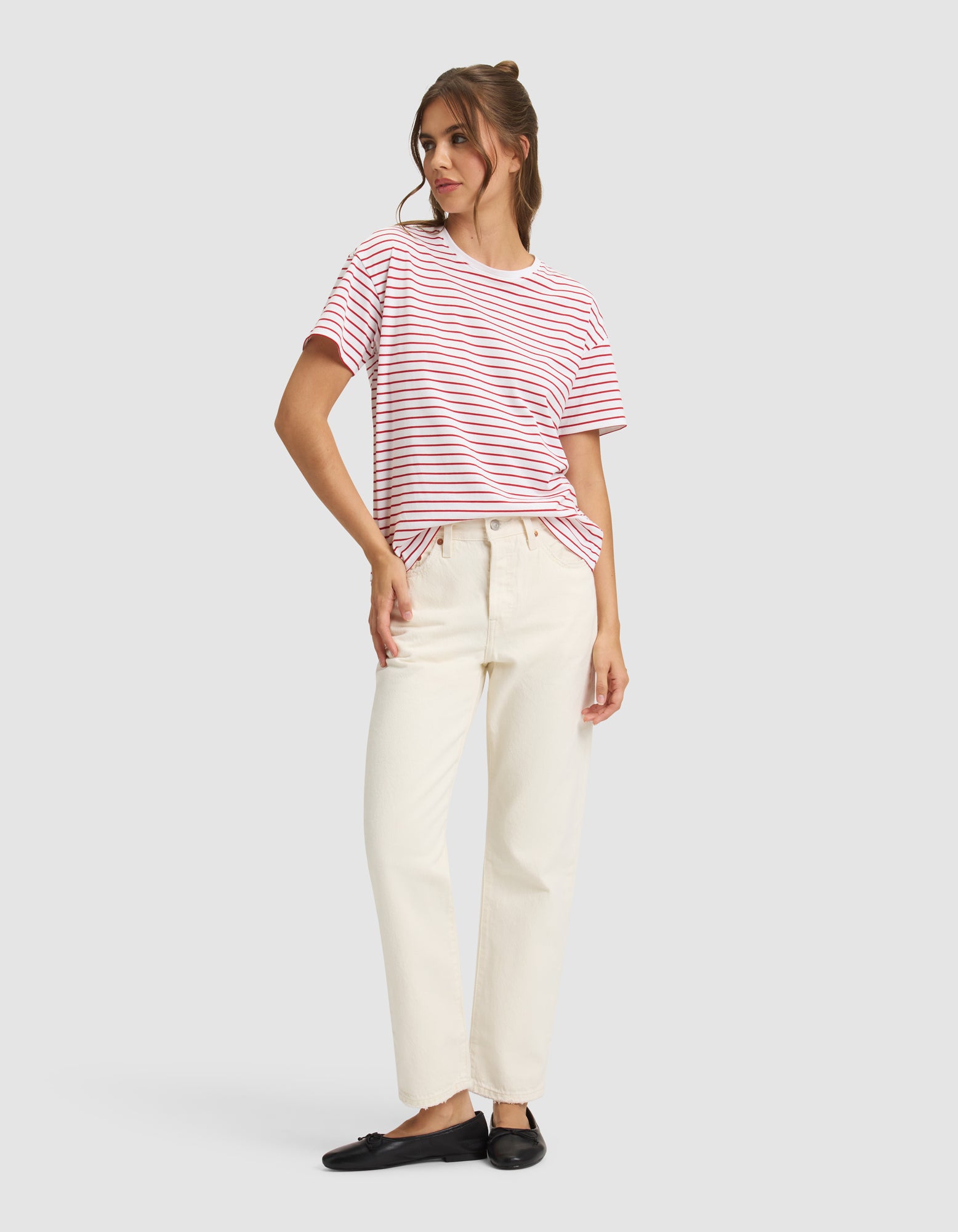 A woman with light brown hair in a half-up bun poses against a light gray background, wearing Cozy Earth's Women's All Day Tee in red and white stripes, paired with cream high-waisted pants and black loafers.