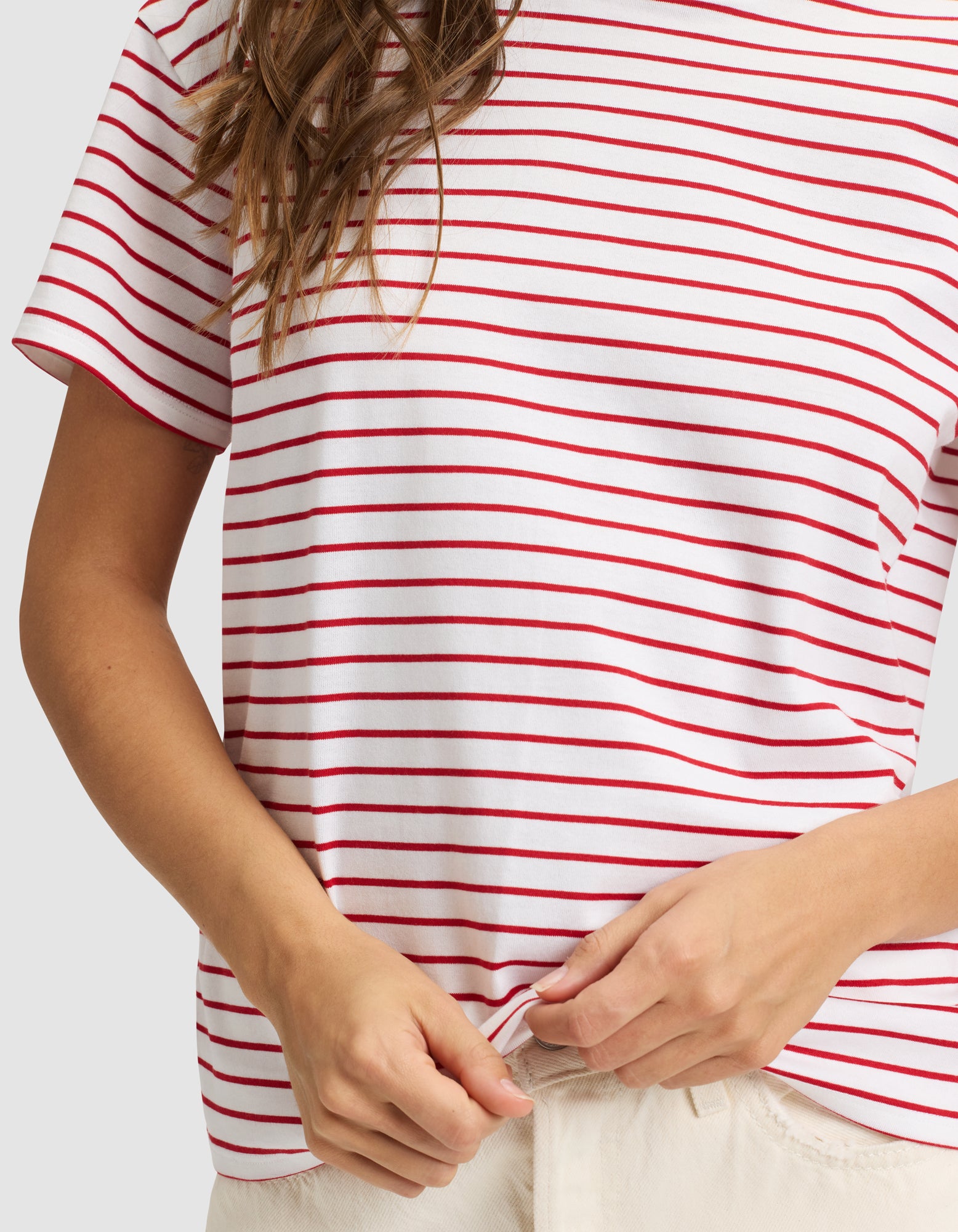 A person models the Cozy Earth Women's All Day Tee—a white shirt with red horizontal stripes—holding the hem with one hand. Only the torso and part of the arms are visible; the face is not shown.