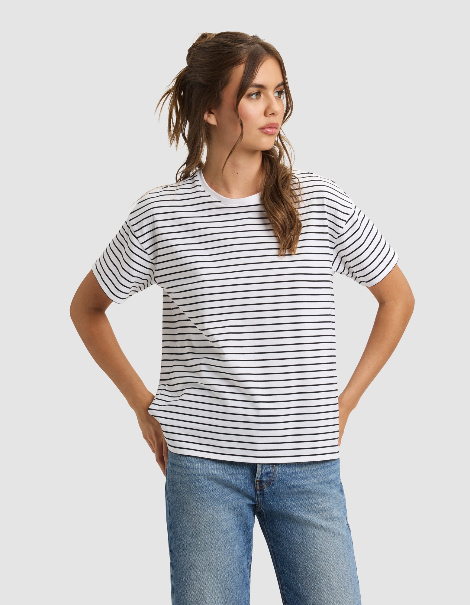 A woman models the Cozy Earth Women's All Day Tee—white with black horizontal stripes—paired with blue jeans, standing hands on hips and looking to the side against a plain light gray background.