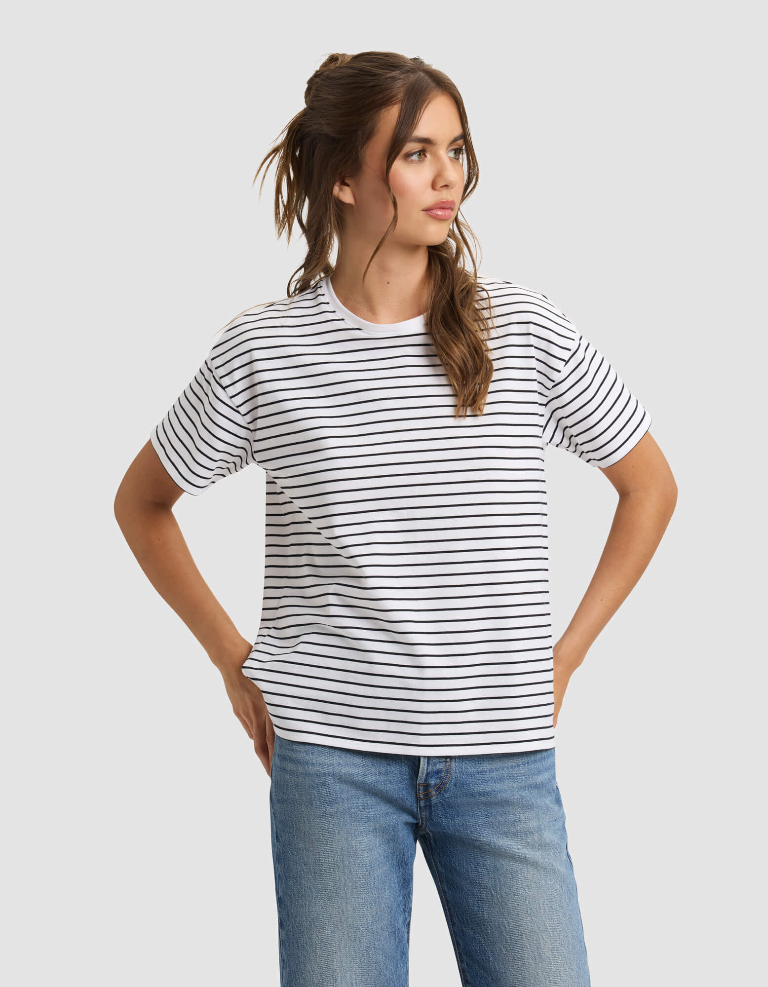A woman models the Cozy Earth Women's All Day Tee—white with black horizontal stripes—paired with blue jeans, standing hands on hips and looking to the side against a plain light gray background. 