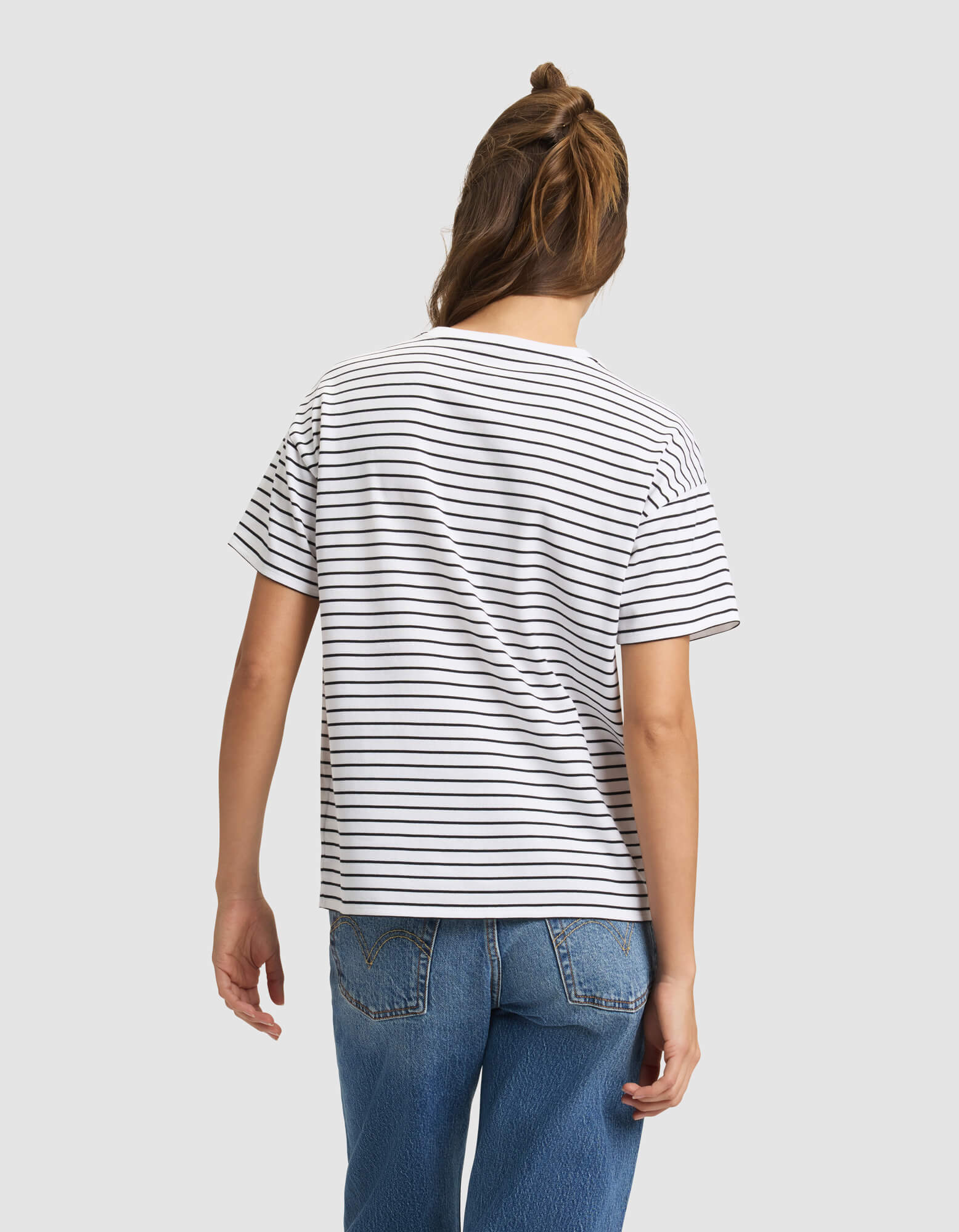 A woman with long brown hair wears the Cozy Earth Women's All Day Tee in white with black stripes and blue jeans, standing with her back to a plain light gray background. 