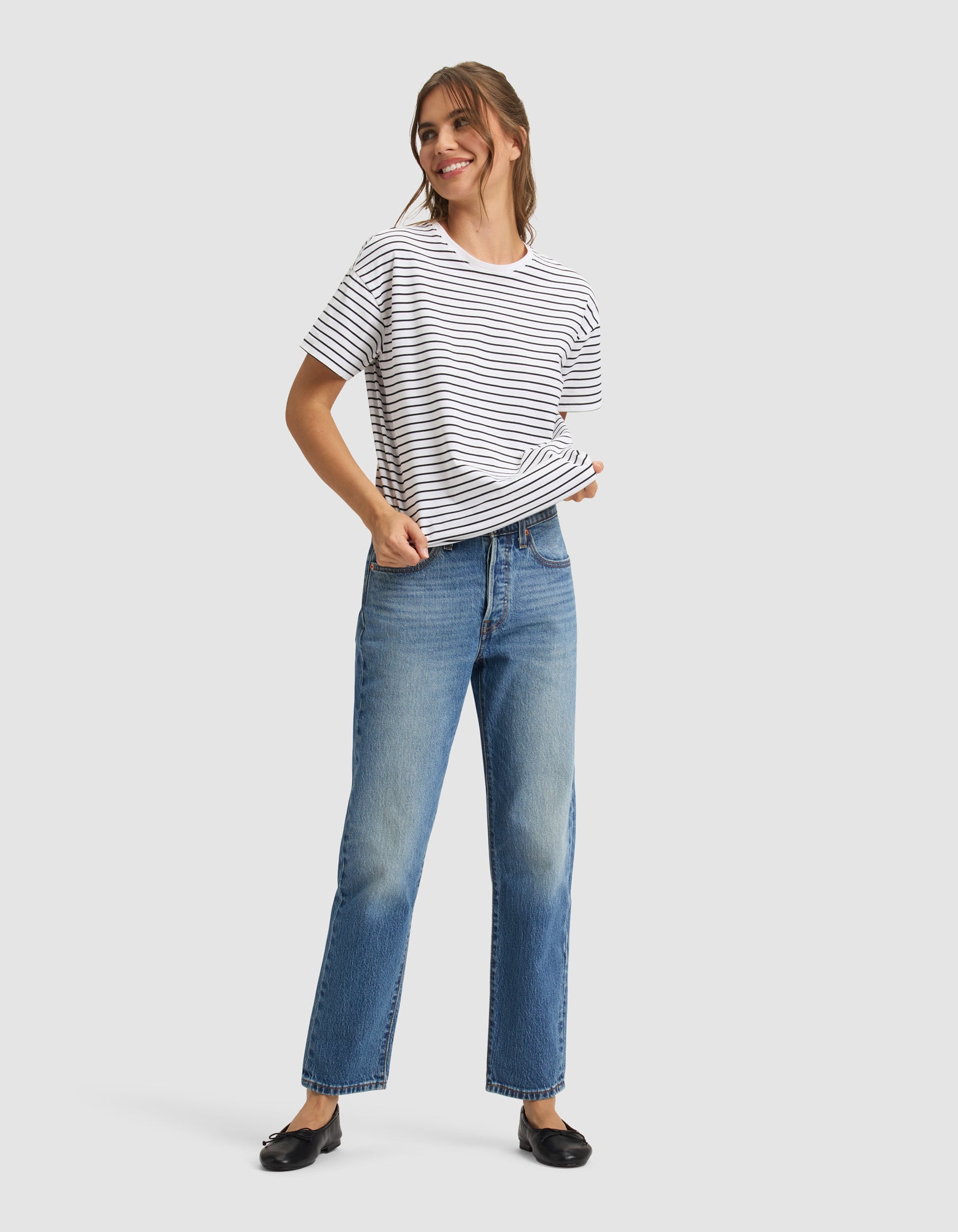 A woman with light brown hair tied back smiles while wearing the Cozy Earth Women's All Day Tee in white and black stripes, paired with blue jeans and black flats. She poses with one hand in her pocket against a plain light gray background. 
