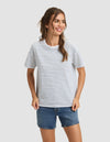 A woman with long brown hair in a ponytail wears the Cozy Earth Women's All Day Tee, featuring thin black stripes, and blue denim shorts. She smiles and looks to the side against a plain light gray background. |Color:Oceanside Stripe