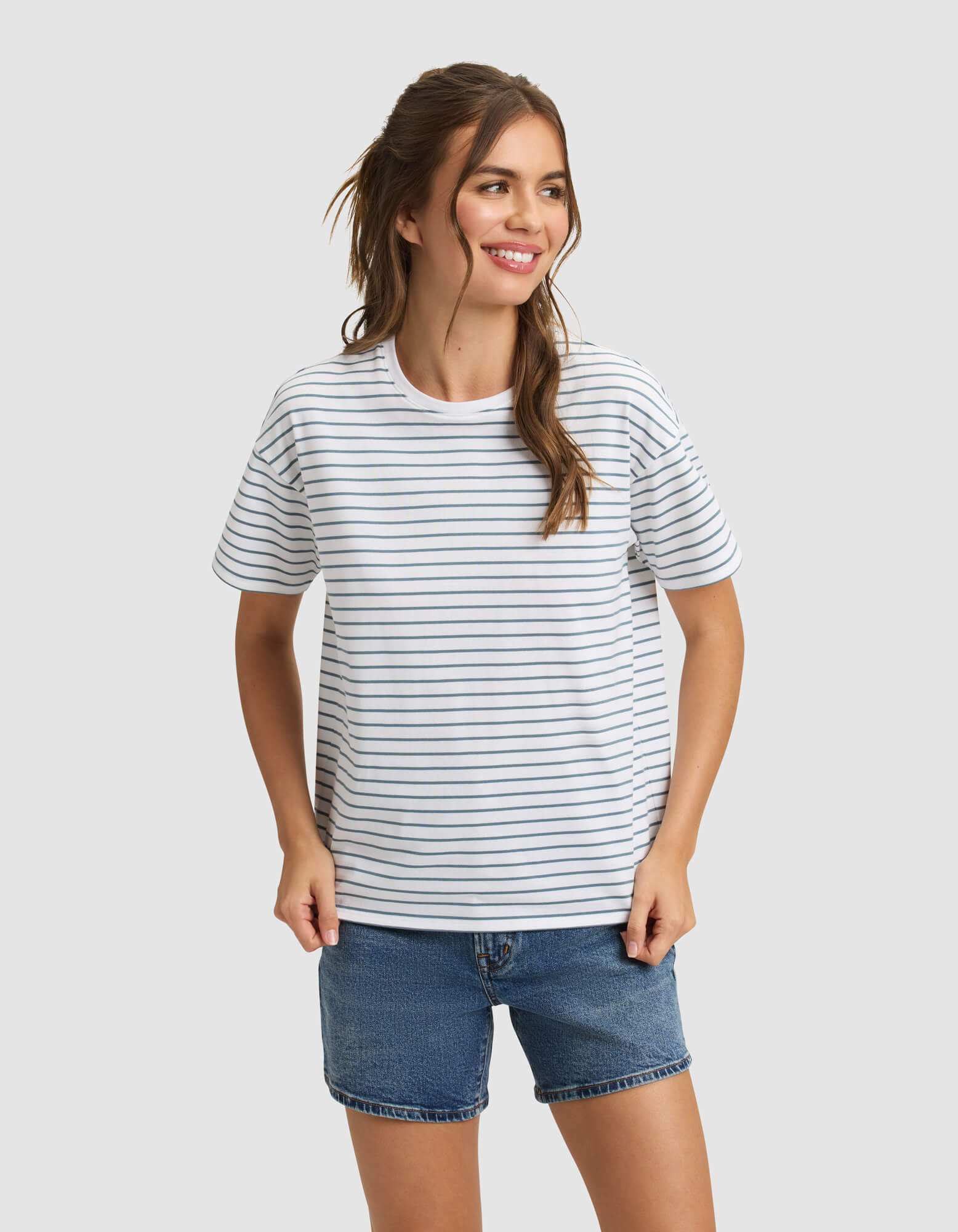 A woman with long brown hair in a ponytail wears the Cozy Earth Women's All Day Tee, featuring thin black stripes, and blue denim shorts. She smiles and looks to the side against a plain light gray background. 