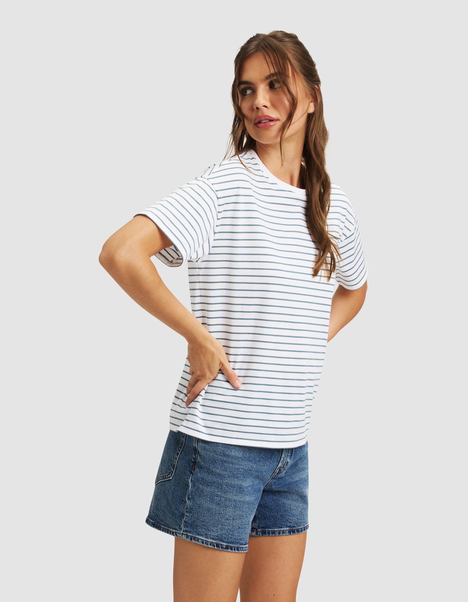 A woman with long brown hair stands hands on hips, looking left, wearing the Cozy Earth Women's All Day Tee—white with thin black stripes—and blue denim shorts against a plain light background.