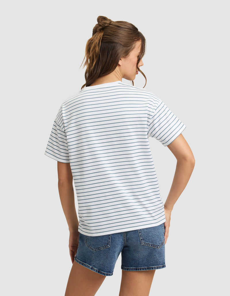 A woman with brown hair faces away, modeling the Cozy Earth Women's All Day Tee—a white T-shirt with thin navy stripes—paired with blue denim shorts against a plain light gray background.