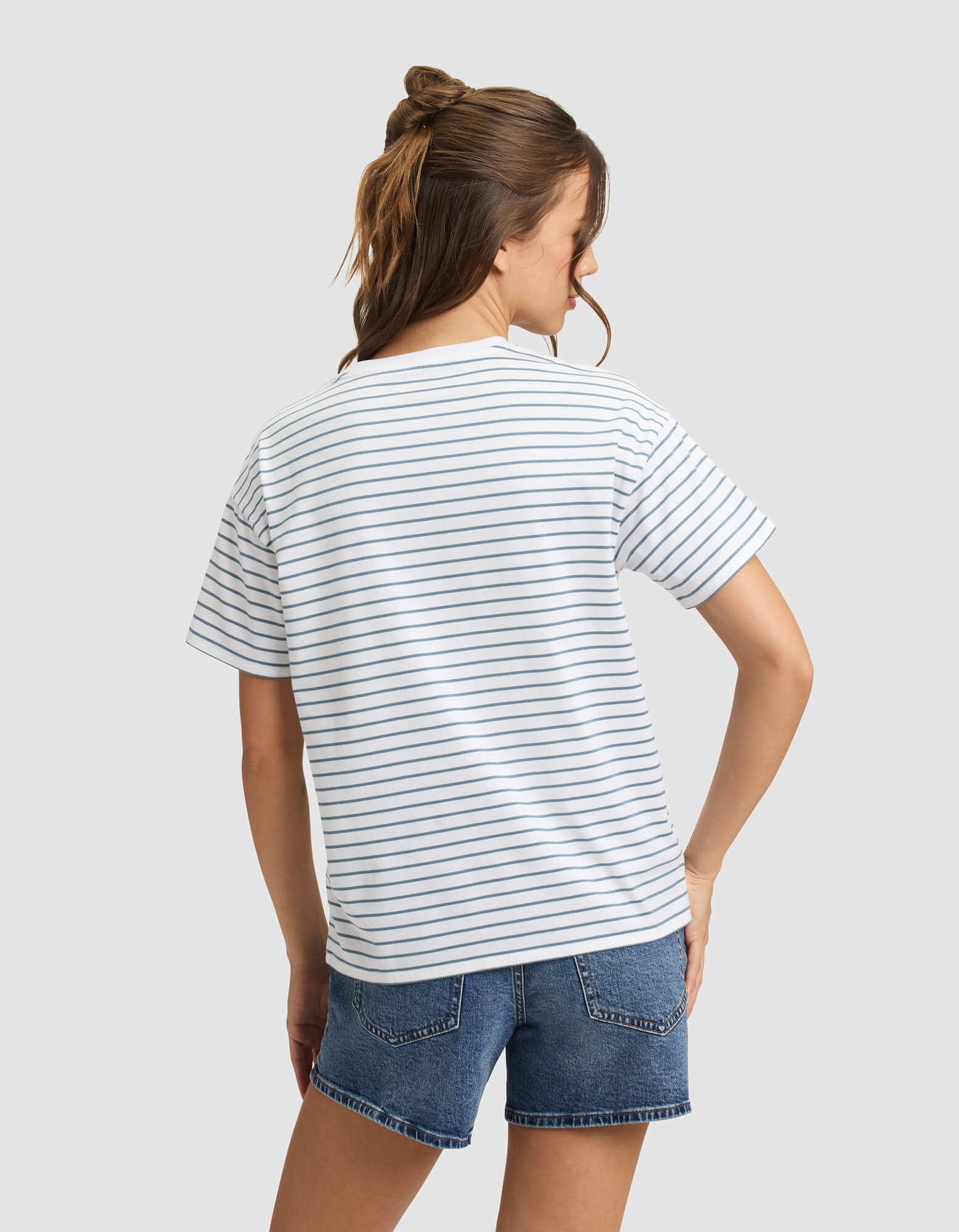 A woman with brown hair faces away, modeling the Cozy Earth Women's All Day Tee—a white T-shirt with thin navy stripes—paired with blue denim shorts against a plain light gray background. 