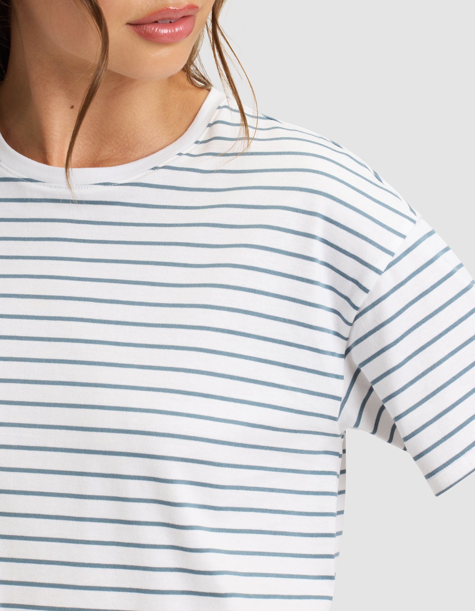 A close-up shows a person wearing Cozy Earth's Women's All Day Tee in white with thin blue horizontal stripes, featuring only the lower face and upper torso against a plain light background.