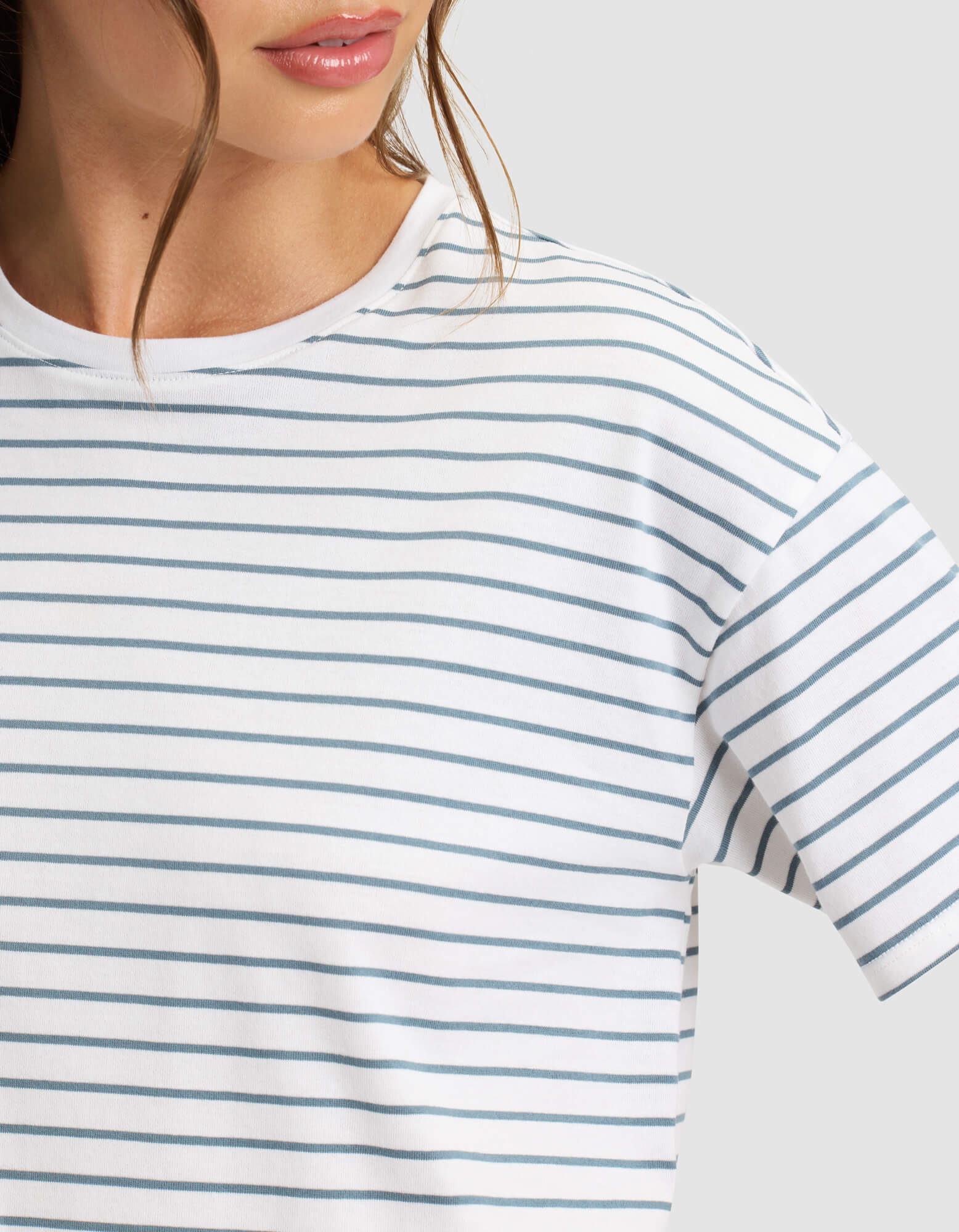 A close-up shows a person wearing Cozy Earth's Women's All Day Tee in white with thin blue horizontal stripes, featuring only the lower face and upper torso against a plain light background. 