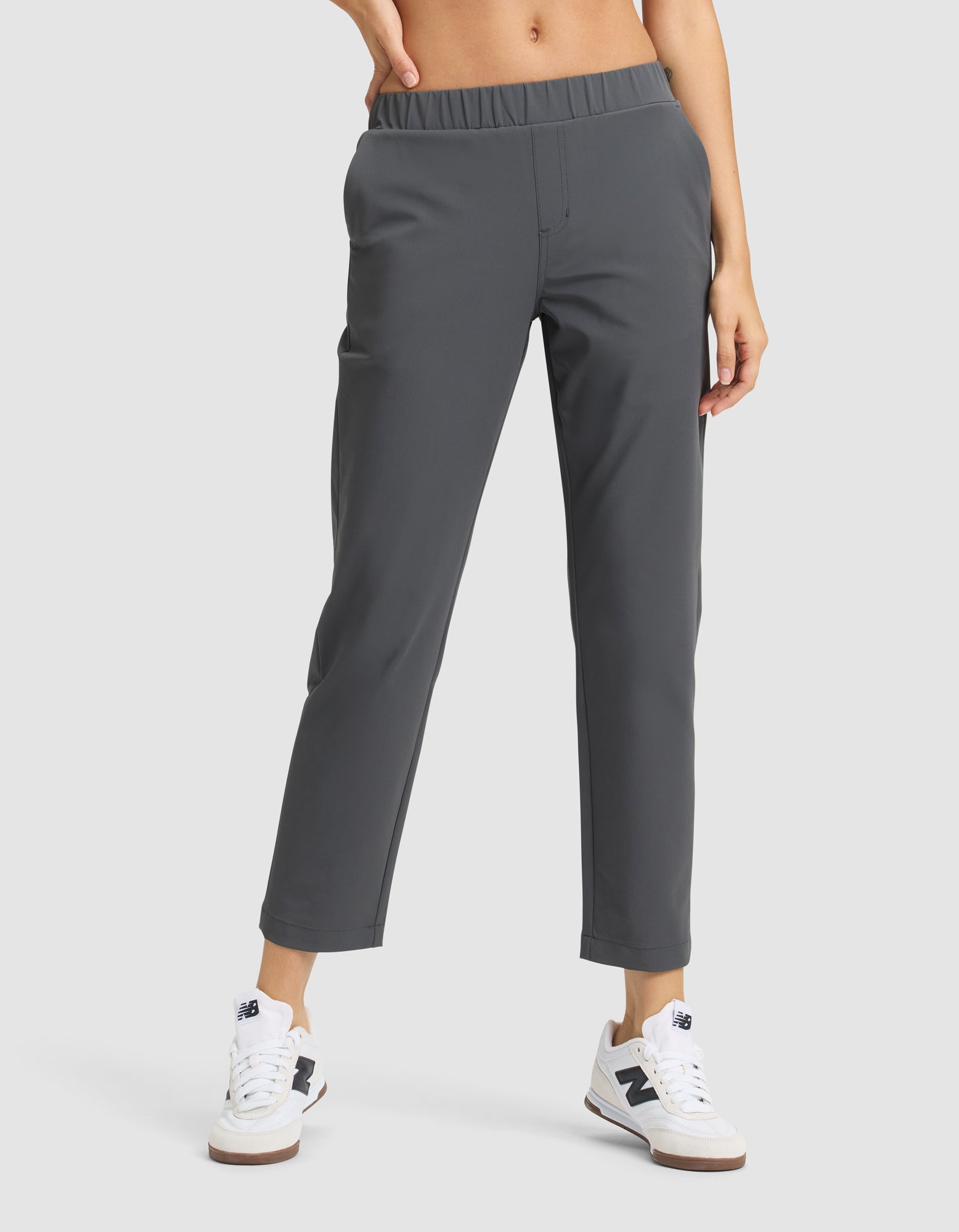 A person wears Cozy Earth’s Women's Always Cropped Pant in dark gray with an elastic waistband, paired with white sneakers featuring blue accents, standing against a plain light background. 