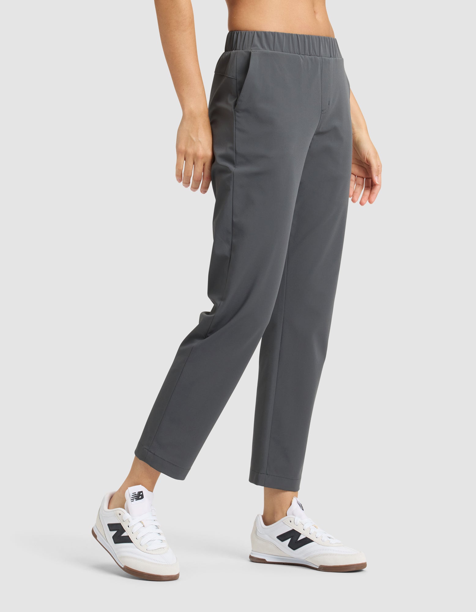 A person wears Cozy Earth's Women's Always Cropped Pant in gray and white New Balance sneakers, standing with one hand in their pocket against a plain light background. 