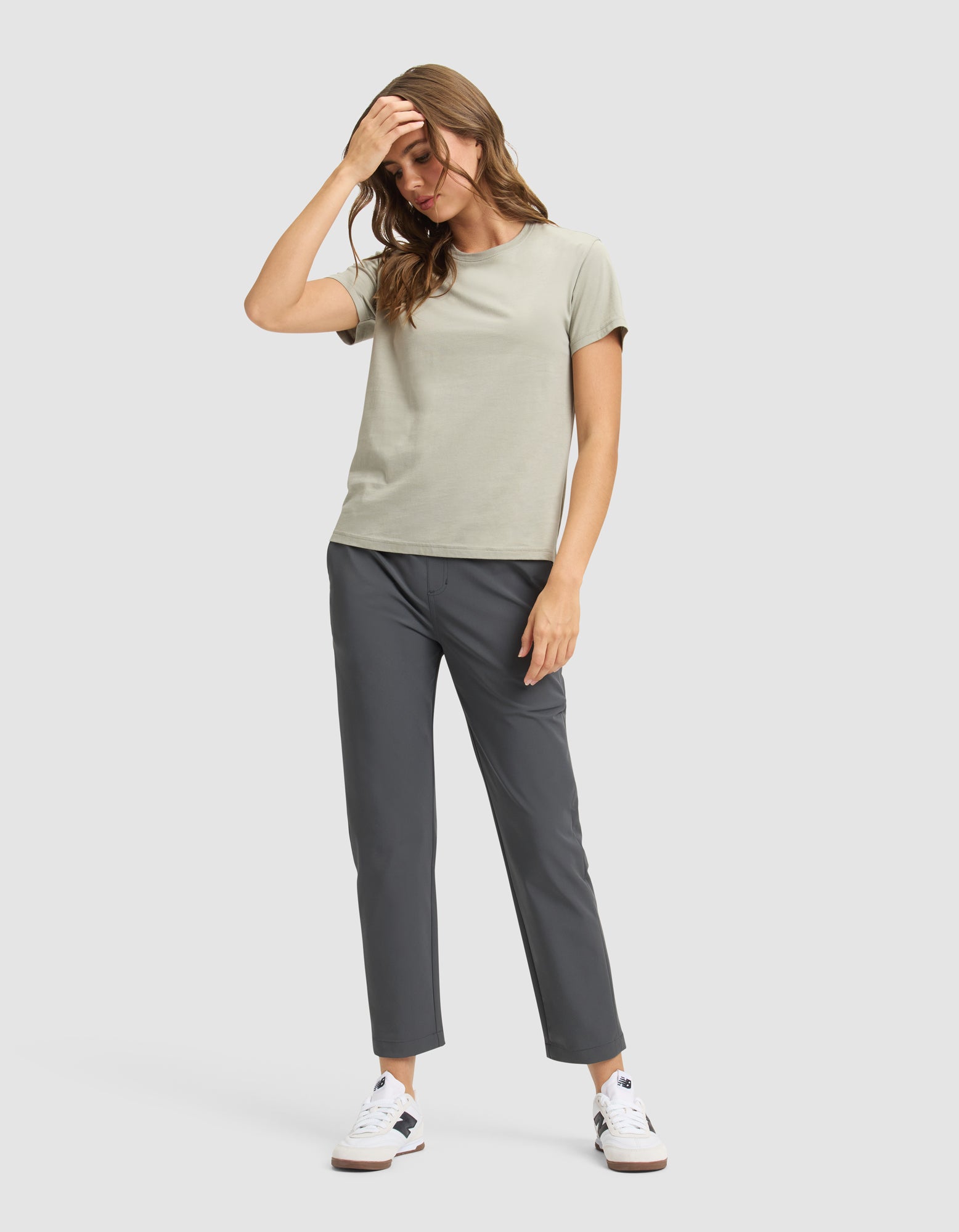 A woman poses casually against a neutral background, wearing Cozy Earth's Women's Always Cropped Pant in dark gray, a light gray t-shirt, and white sneakers. She has one hand on her head and her hair is loose for a relaxed look. 