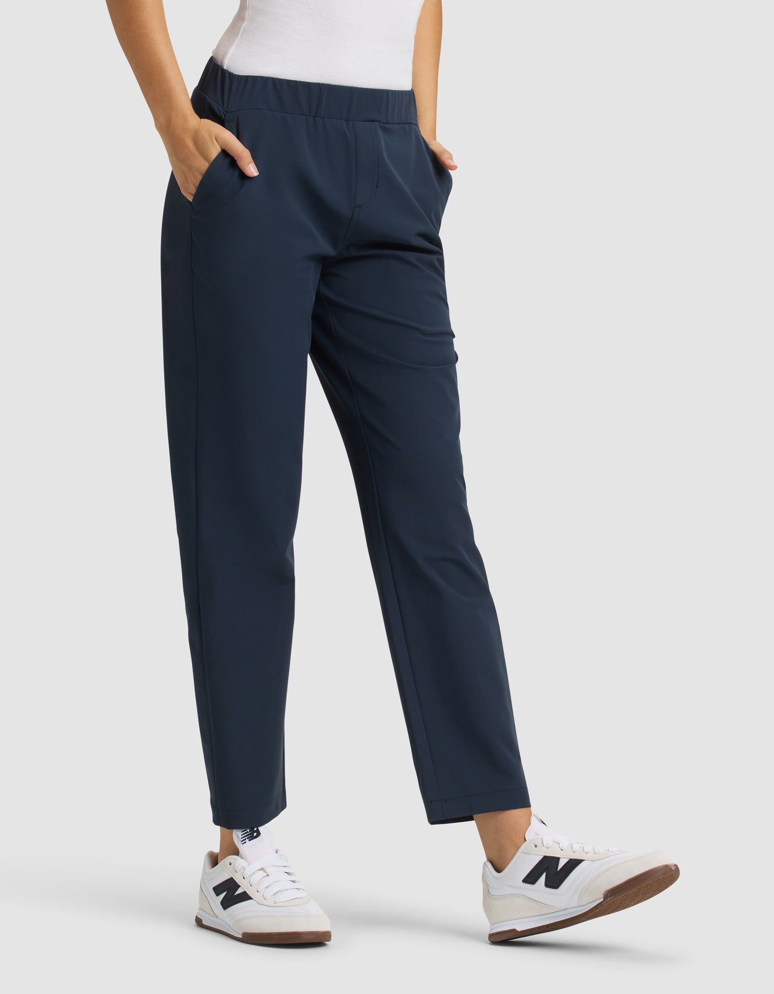 A person wears Cozy Earth Women's Always Cropped Pant in navy blue with a white top and white sneakers with brown soles. Hands are in the pants' pockets, standing against a plain light gray background. 