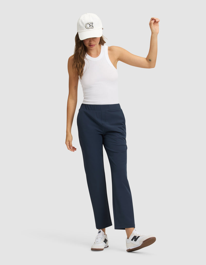 A woman models the Cozy Earth Women's Always Cropped Pant in navy blue, paired with a white sleeveless tank, white sneakers, and a white cap, standing against a light gray background with one arm raised and the other at her side.