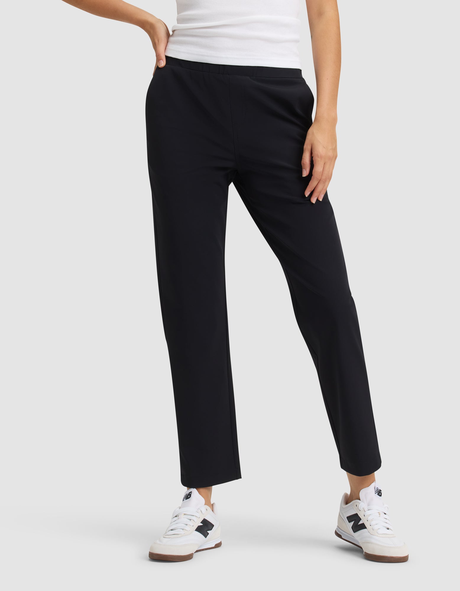 Wearing a white tank top, Cozy Earth Women's Always Cropped Pant in black, and white New Balance sneakers, a person stands with one hand on their hip against a plain background. 