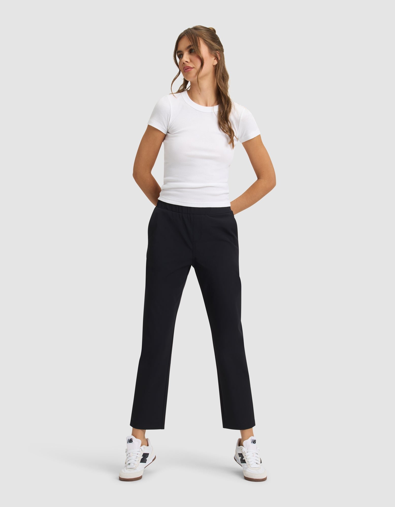 A woman stands against a light gray background, smiling with her hands in her pockets, wearing Cozy Earth's Women's Always Cropped Pant in black, a fitted white t-shirt, and white sneakers with blue and tan accents. 