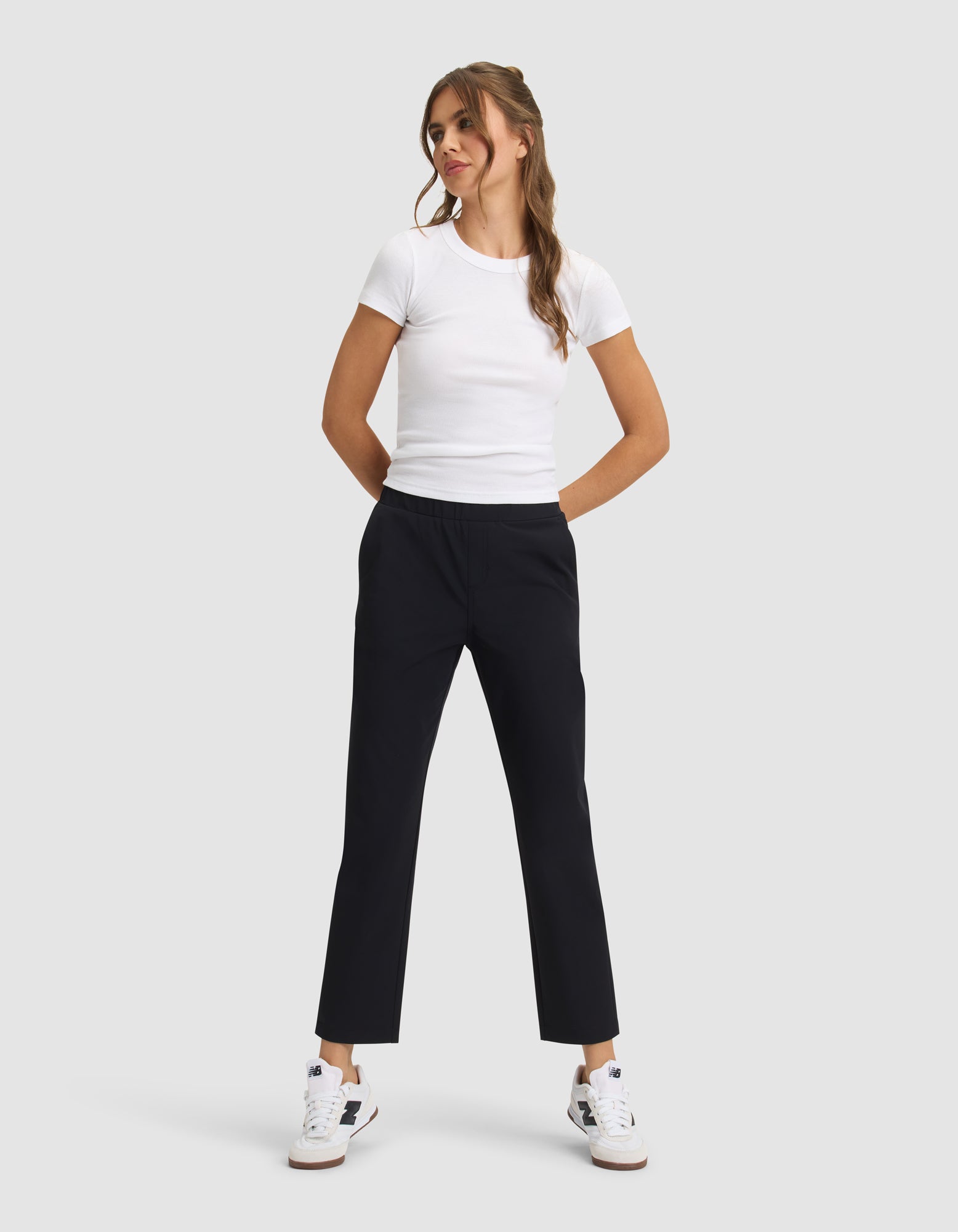 A woman stands against a light gray background, smiling with her hands in her pockets, wearing Cozy Earth's Women's Always Cropped Pant in black, a fitted white t-shirt, and white sneakers with blue and tan accents. 