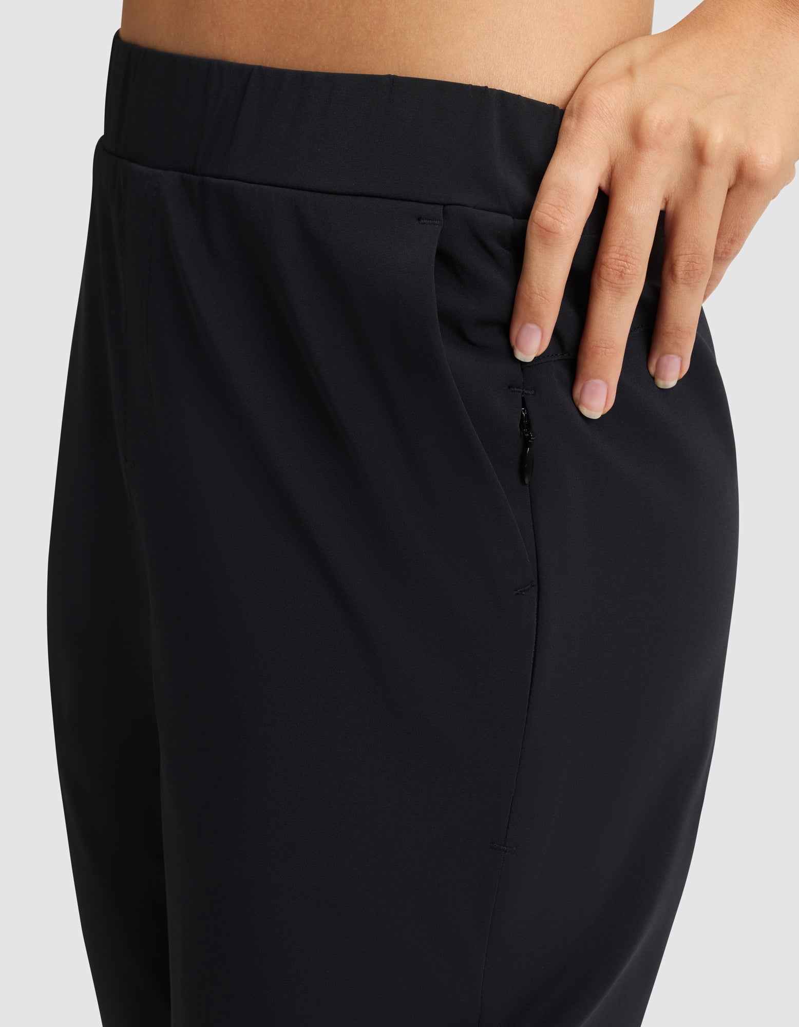A close-up of a person wearing Cozy Earth's Women's Always Cropped Pant in black, featuring a zipper pocket and their hand resting on the waistband against a plain, light-colored background. 