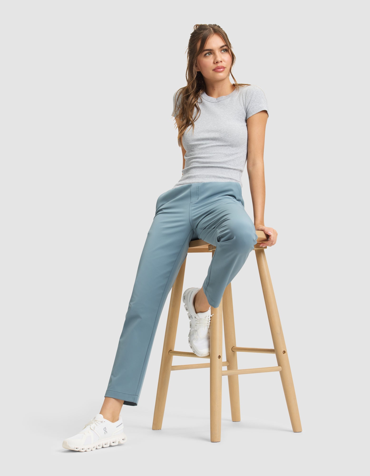 A woman in a grey t-shirt and white sneakers wears Cozy Earth's Women's Always Cropped Pant in light blue, sitting casually on a wooden stool against a plain, light grey background. 