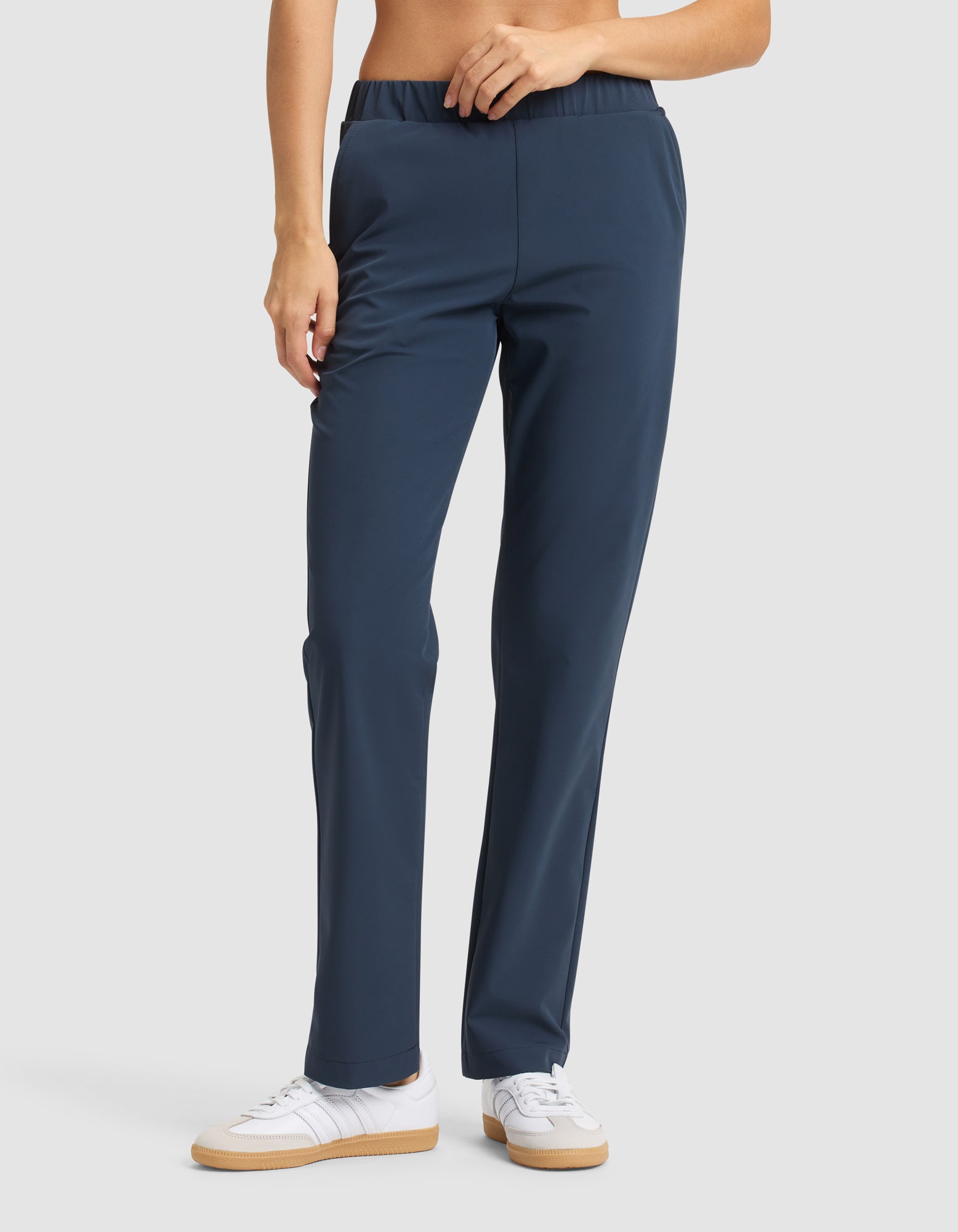 A woman in Cozy Earth's Women's Always Pant, navy blue with a straight-leg cut and elastic waistband, paired with white sneakers, stands against a plain light background. 