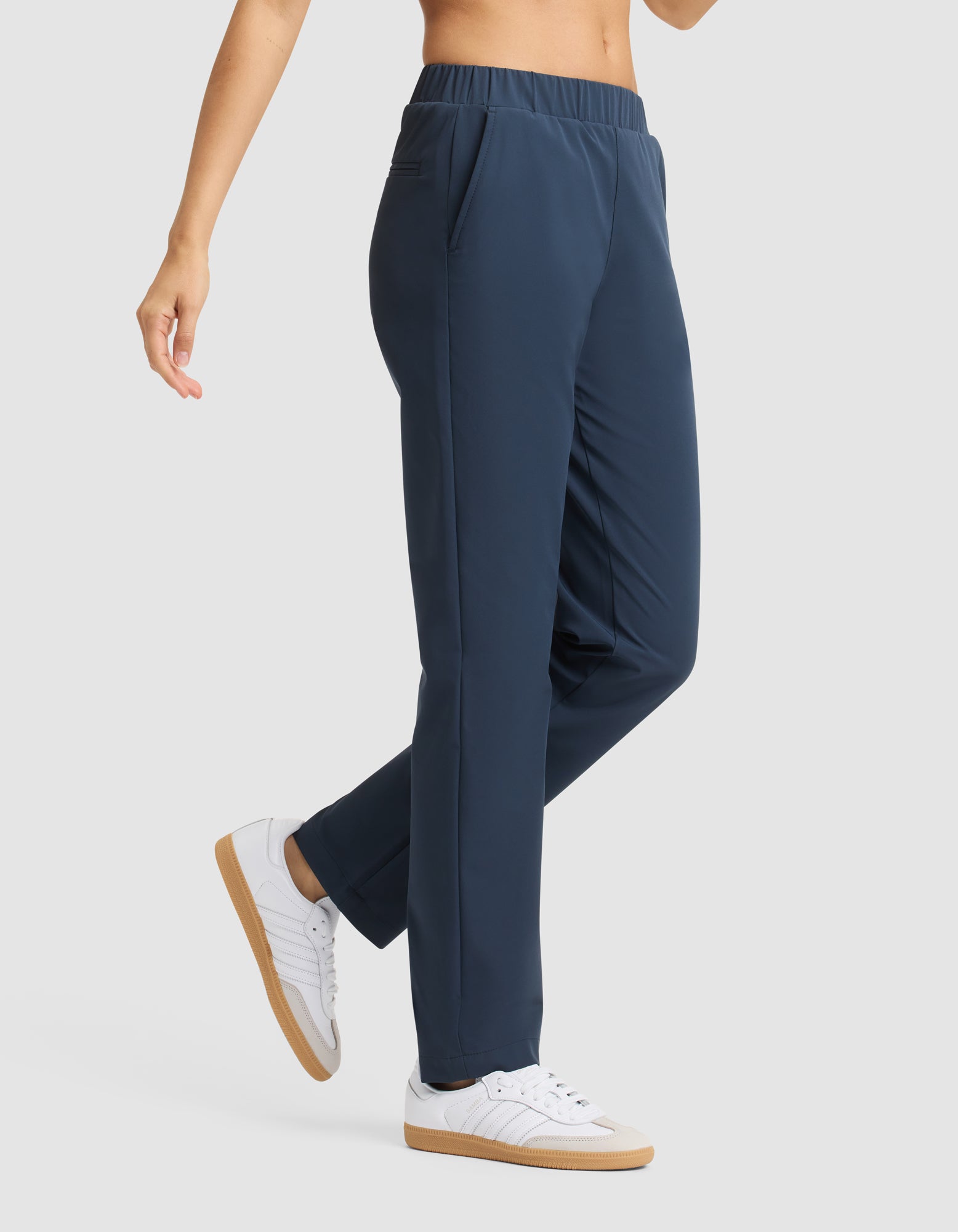 A woman models Cozy Earth's Women's Always Pant in navy blue, featuring an elastic waistband and pockets, paired with white sneakers. Only her lower torso and legs are visible against a light gray background. 