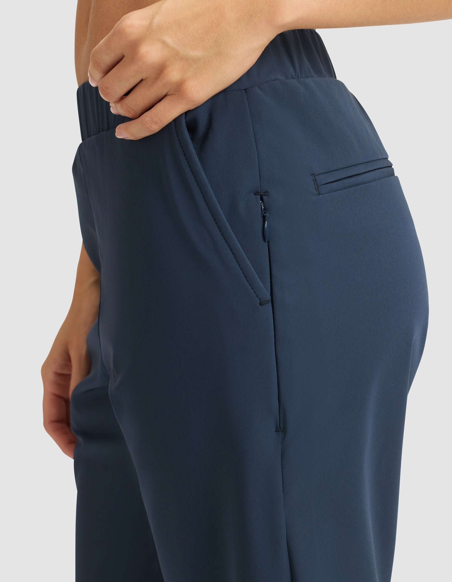 A close-up of a person wearing Cozy Earth's Women's Always Pant in dark blue with an elastic waistband. One hand is near the zippered side pocket. The background is plain and light-colored. 