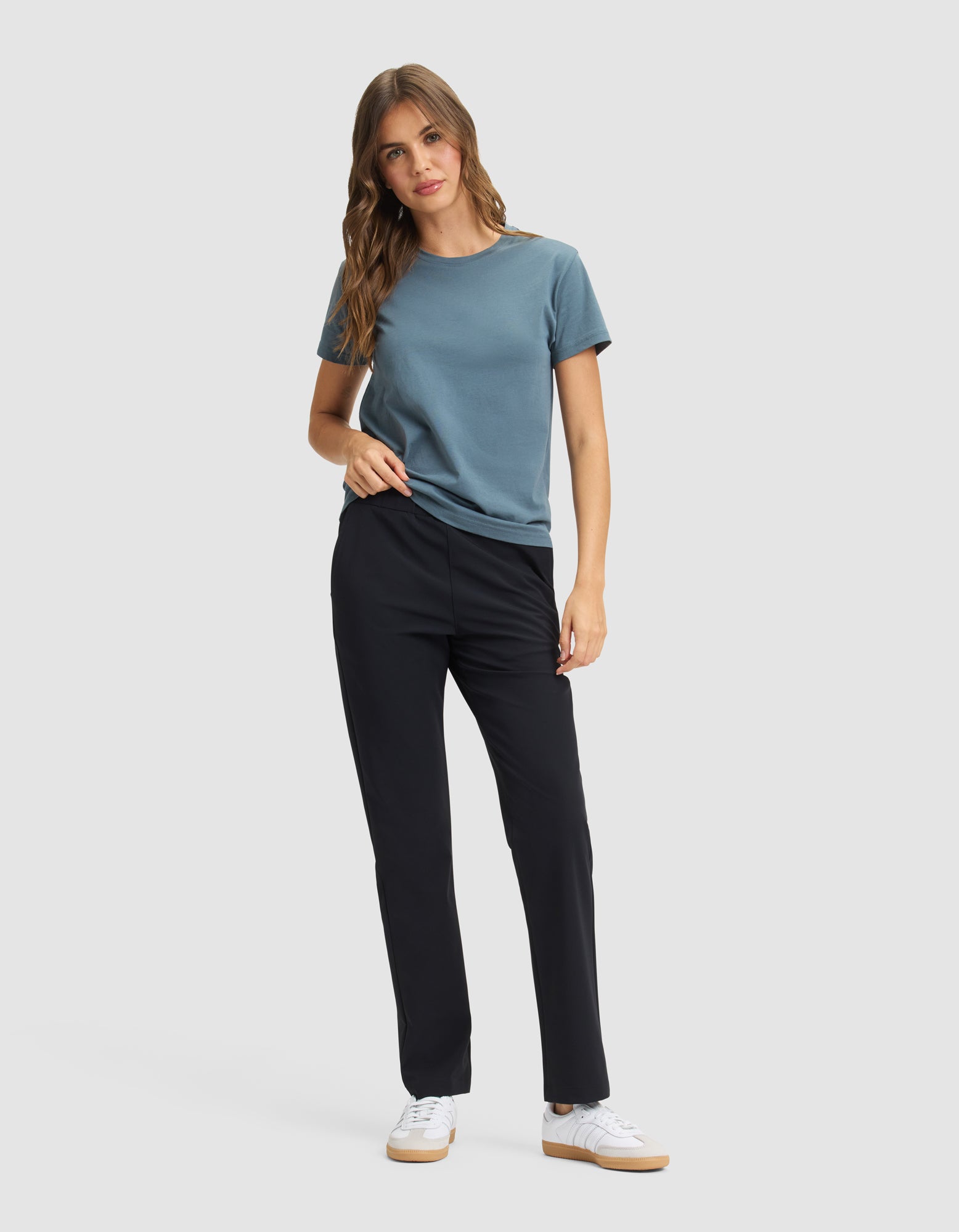 A woman with long, wavy hair poses against a plain background, wearing Cozy Earth's Women's Always Pant in black, paired with a blue short-sleeve t-shirt and white sneakers. One hand is in her pocket. 