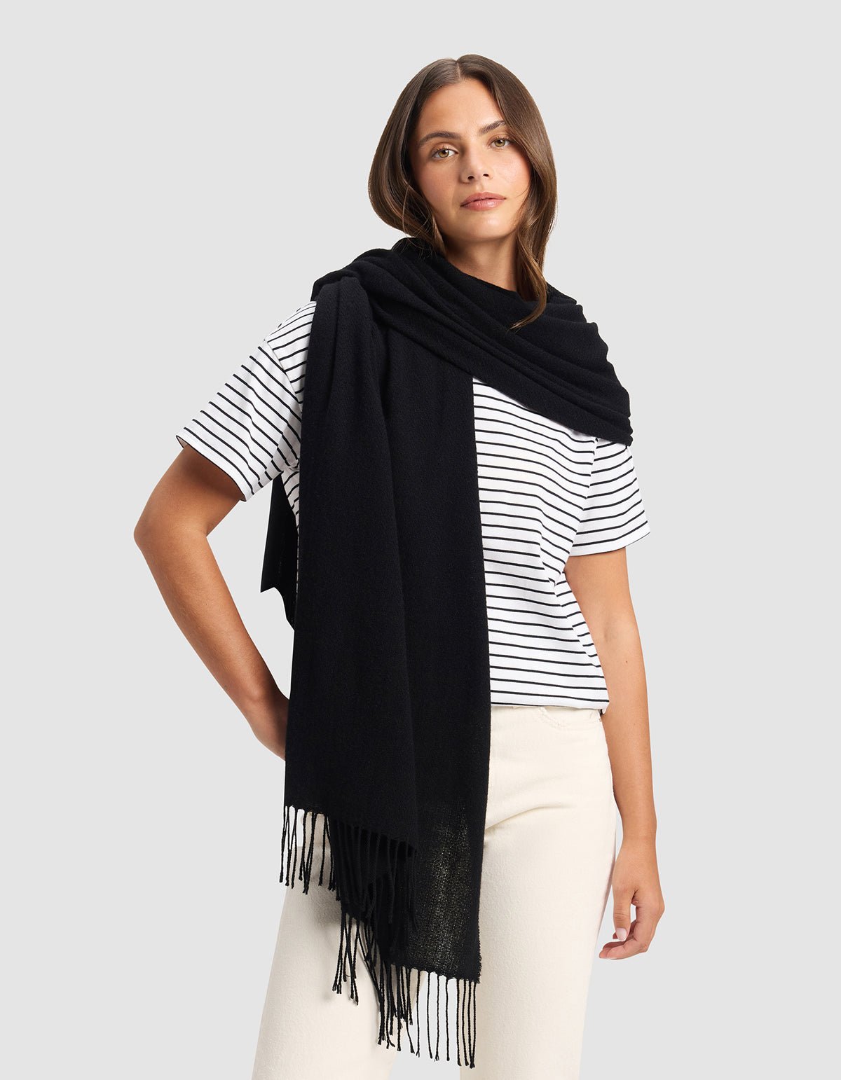 A woman stands against a plain gray background wearing the Cozy Earth Amelia Wrap in black over her shoulders, paired with a white and black striped t-shirt and cream-colored pants.