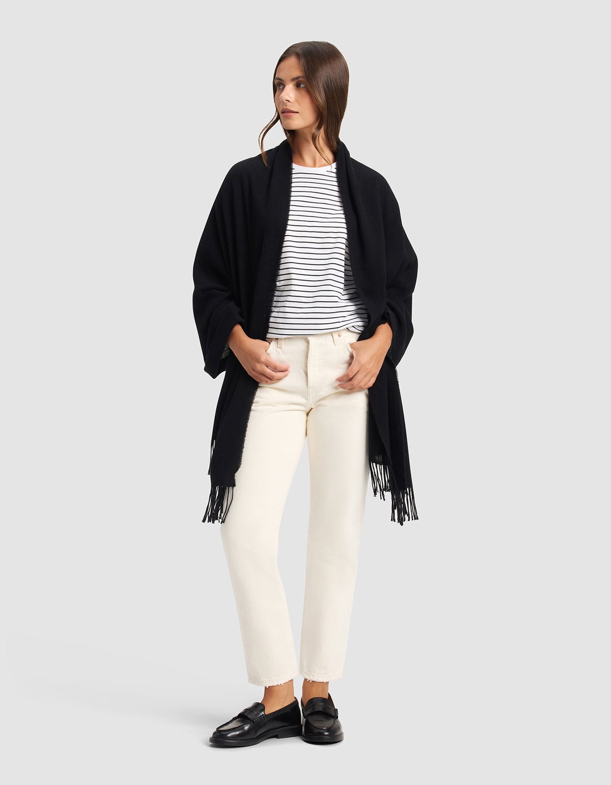 A woman stands against a plain background, wearing the Cozy Earth Amelia Wrap—a black shawl with fringe—over a white and black striped shirt, off-white pants, and black loafers. She looks to the side with her hands in her pockets.