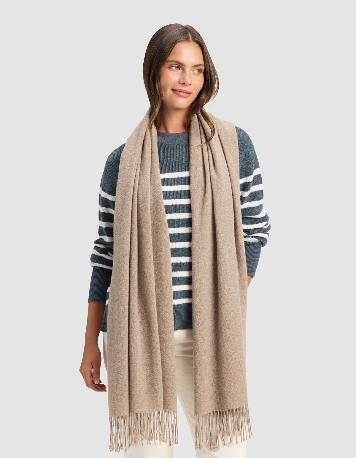 A woman stands against a light gray background wearing the Cozy Earth Amelia Wrap, a long beige scarf with fringed ends, paired with a blue and white striped sweater and cream-colored pants. 