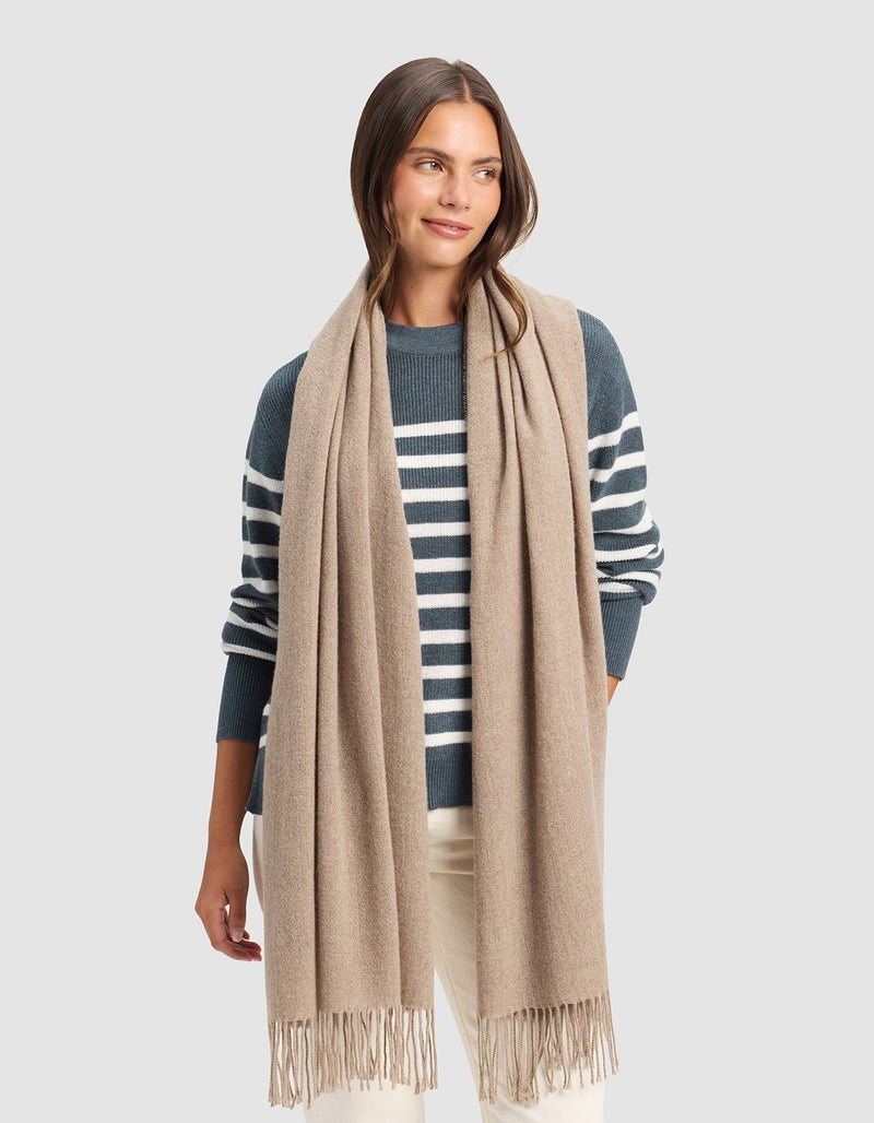 A woman stands against a light gray background wearing the Cozy Earth Amelia Wrap, a long beige scarf with fringed ends, paired with a blue and white striped sweater and cream-colored pants.