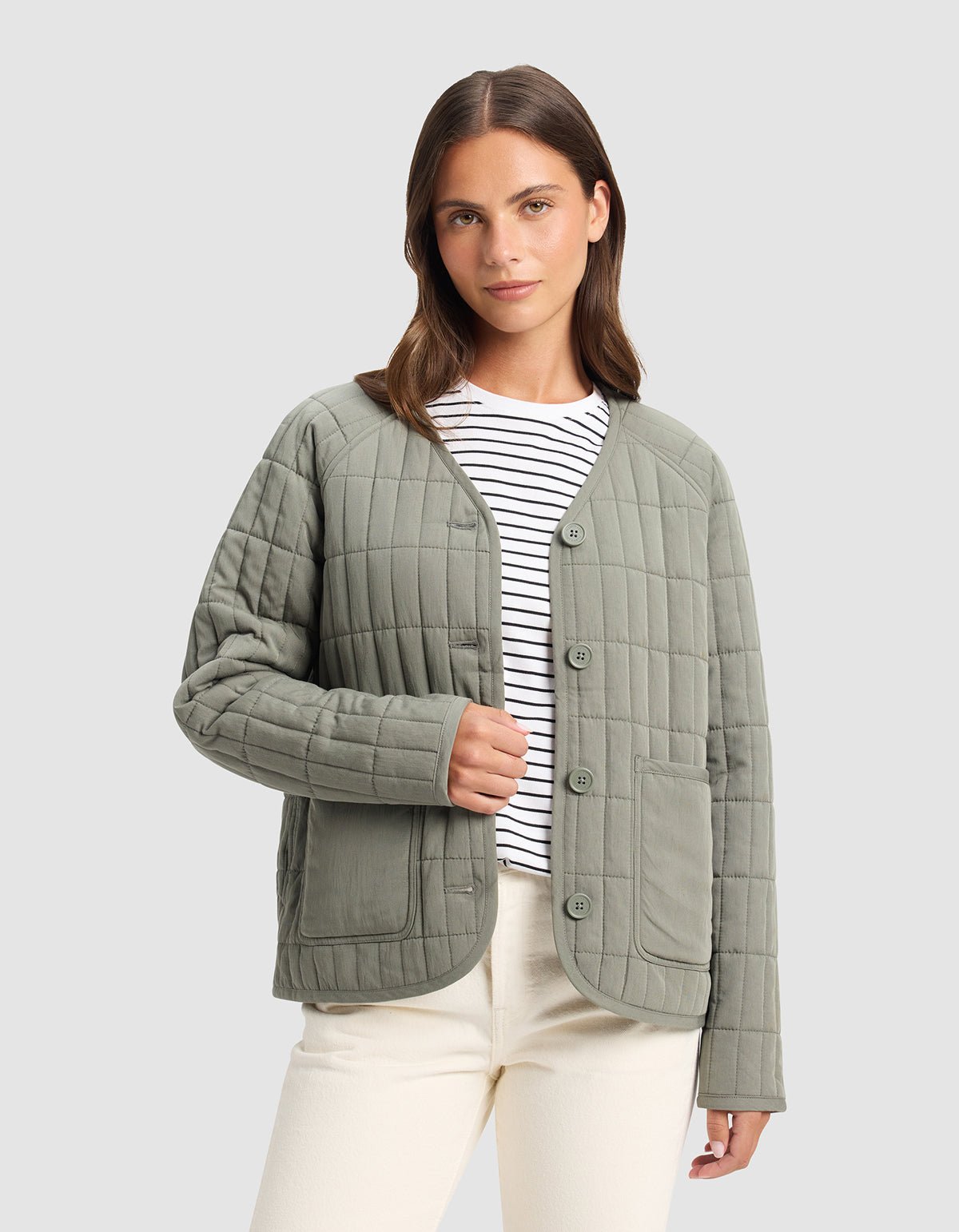 A woman wears the Cozy Earth Women's Andie Quilted Jacket in light green over a white and black striped shirt with cream-colored pants, standing against a plain light gray background.|Color:Cypress