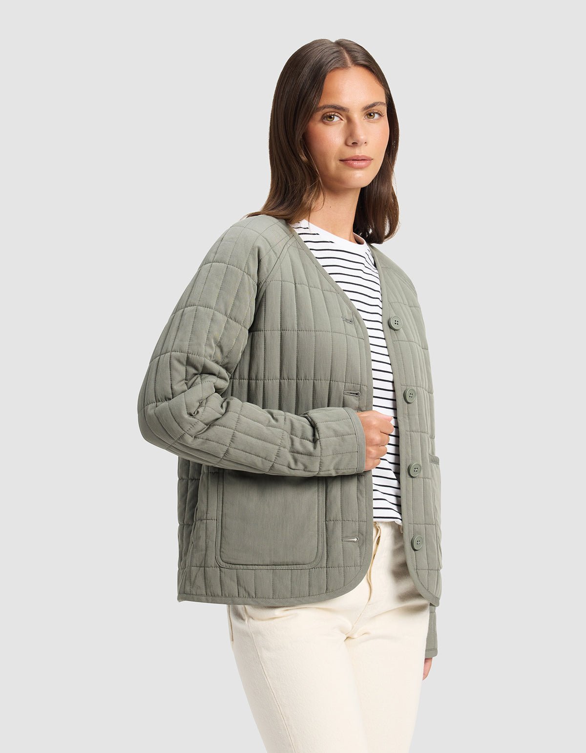A woman wears the Cozy Earth Women's Andie Quilted Jacket in light olive green over a black-and-white striped shirt and cream pants, standing against a plain gray background.
