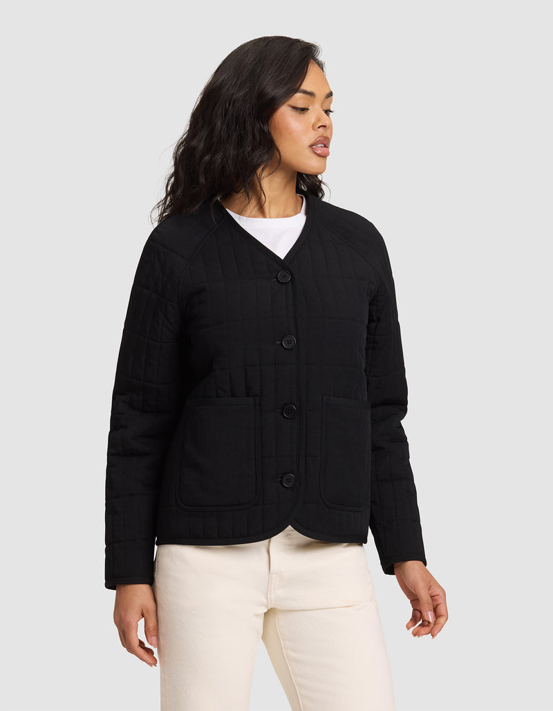 A woman stands against a plain background wearing the Cozy Earth Women's Andie Quilted Jacket in black, paired with a white shirt and off-white pants. She looks to the side with her arms relaxed at her sides.