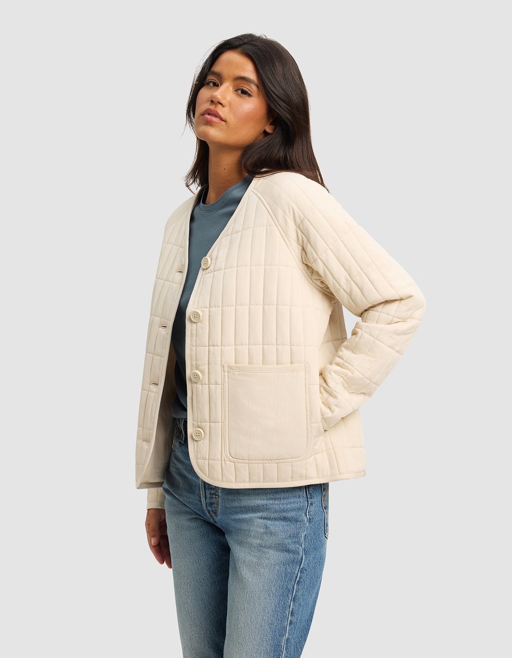 A woman with long dark hair wears the Cozy Earth Women's Andie Quilted Jacket in cream over a blue top and light blue jeans, standing with one hand in her pocket against a plain light gray background.|Color:Sesame