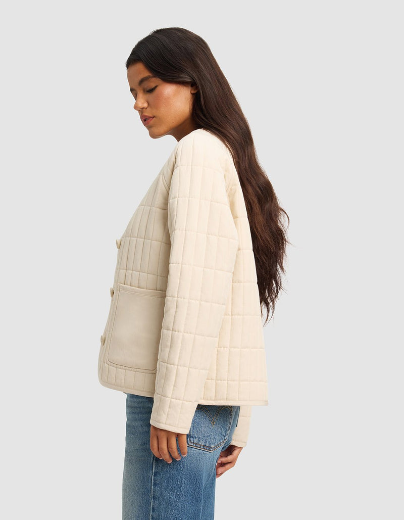 A woman with long dark hair stands in profile wearing the Cozy Earth Women's Andie Quilted Jacket in cream, featuring large front pockets, paired with blue jeans against a plain light gray background.