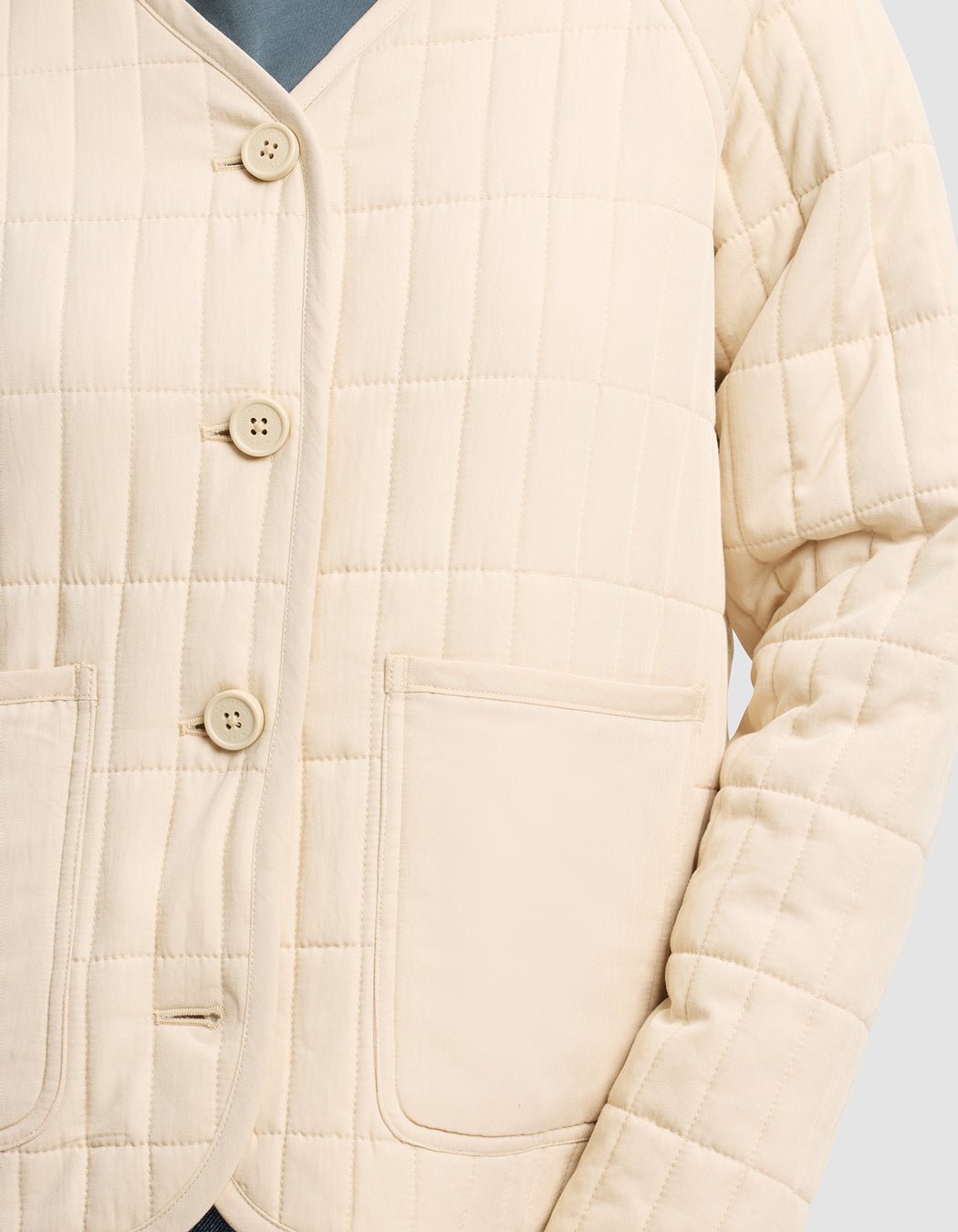 A close-up of the Women's Andie Quilted Jacket by Cozy Earth, shown in cream with grid pattern stitching, four buttons, and two front pockets, worn over a blue top.