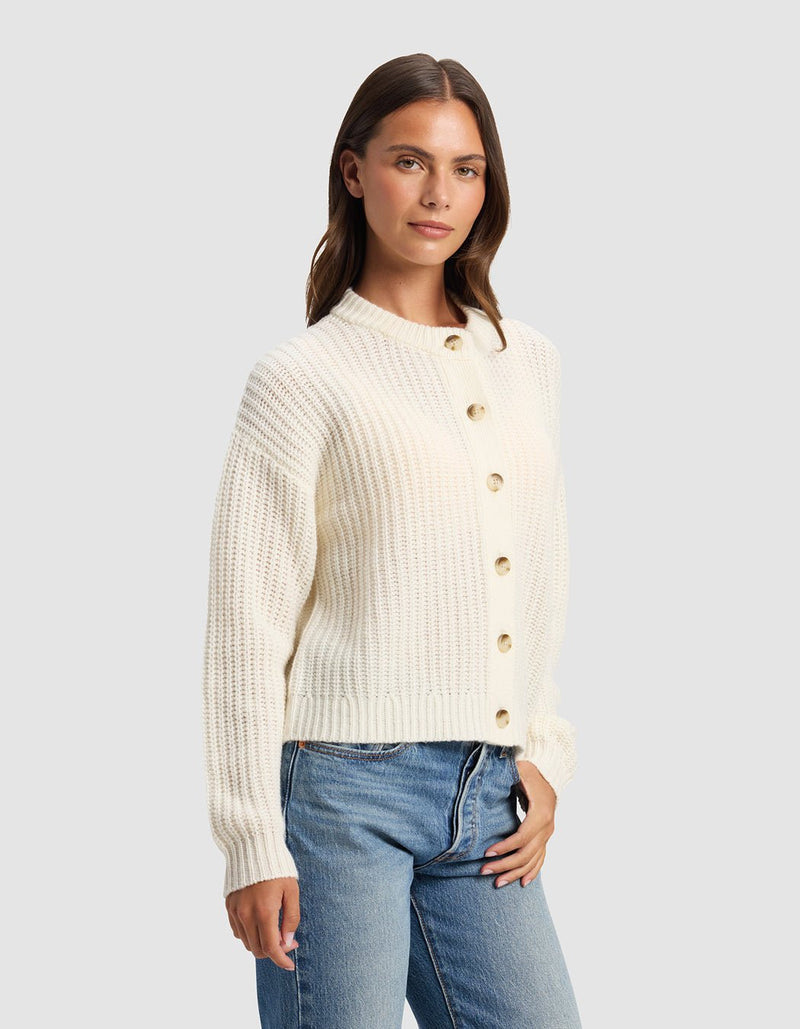 A woman wearing the Cozy Earth Women's Arden Cardigan, a cream-colored ribbed button-up sweater with long sleeves, styled with blue jeans and standing against a plain light gray background.