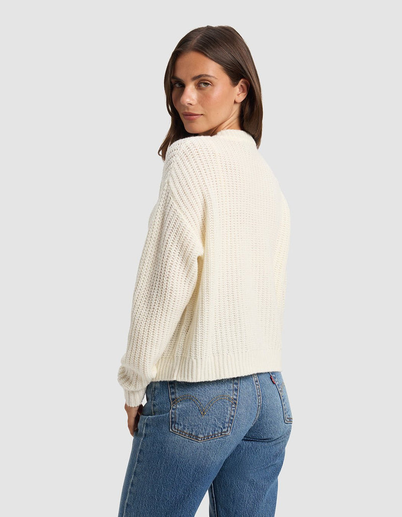 A woman with straight brown hair models the Cozy Earth Women's Arden Cardigan in off-white, paired with blue jeans, as she stands sideways and glances over her shoulder against a plain light gray background.