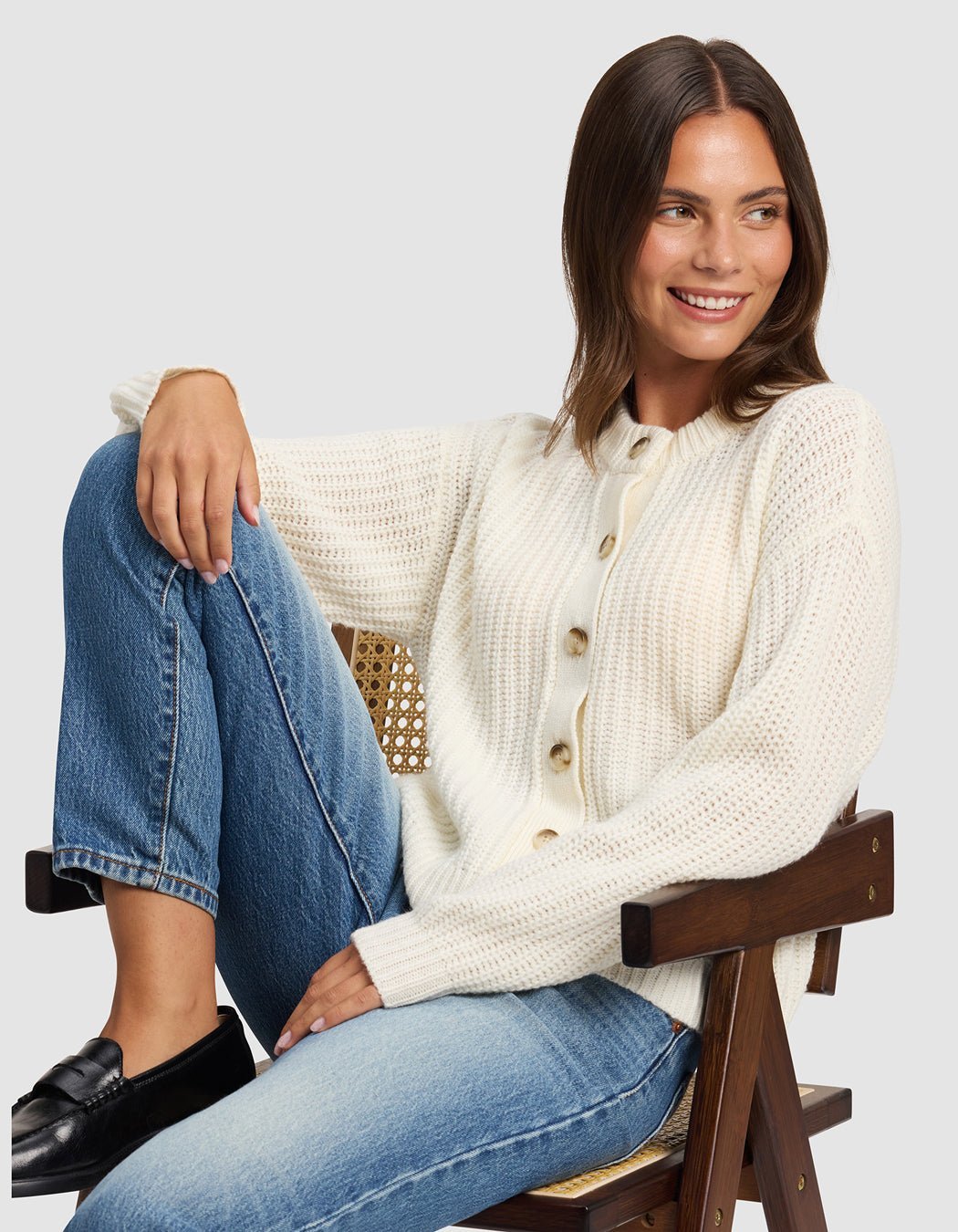 A woman with brown hair smiles as she sits sideways on a wooden chair, wearing Cozy Earth's Women's Arden Cardigan in cream, blue jeans, and black loafers. One leg is bent on the seat against a plain light gray background.